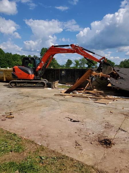 An orange excavator demolishing a small building structure outdoors under a partly cloudy blue sky.