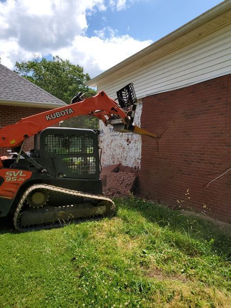 An orange Kubota skid steer with a demolition attachment removing a section of red brick wall on a house.