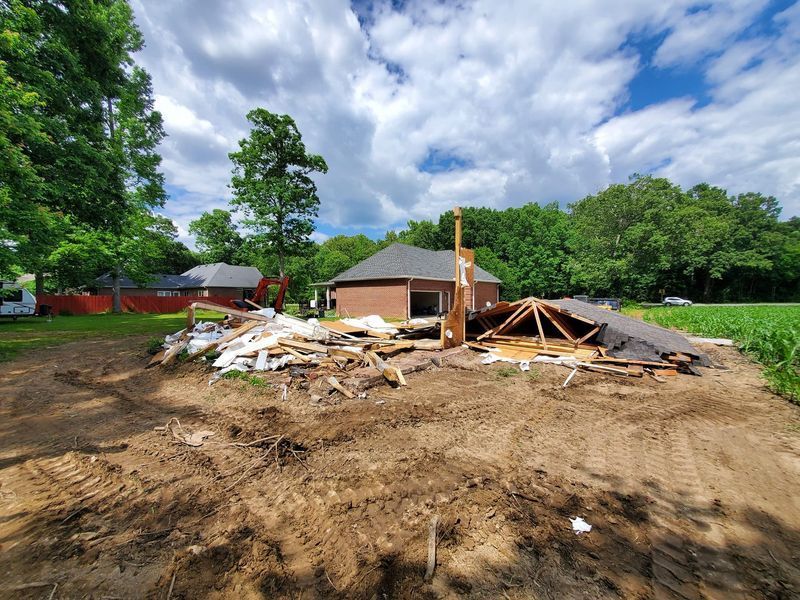 Debris from a demolished building sits on a dirt lot with a partially standing brick house and trees in the background.
