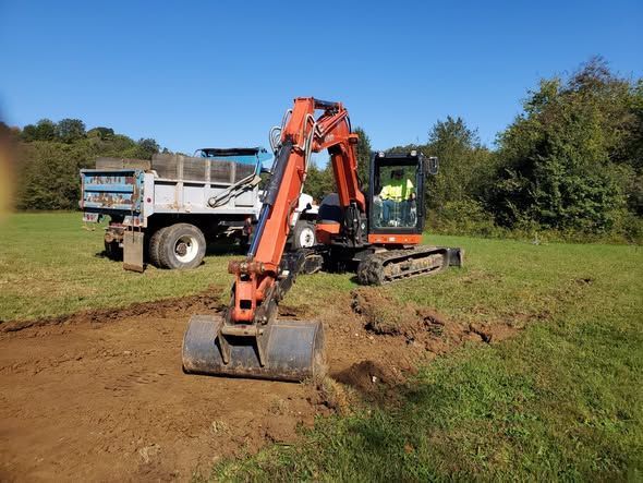 An orange excavator digs into a grassy field next to a dump truck under a clear blue sky.