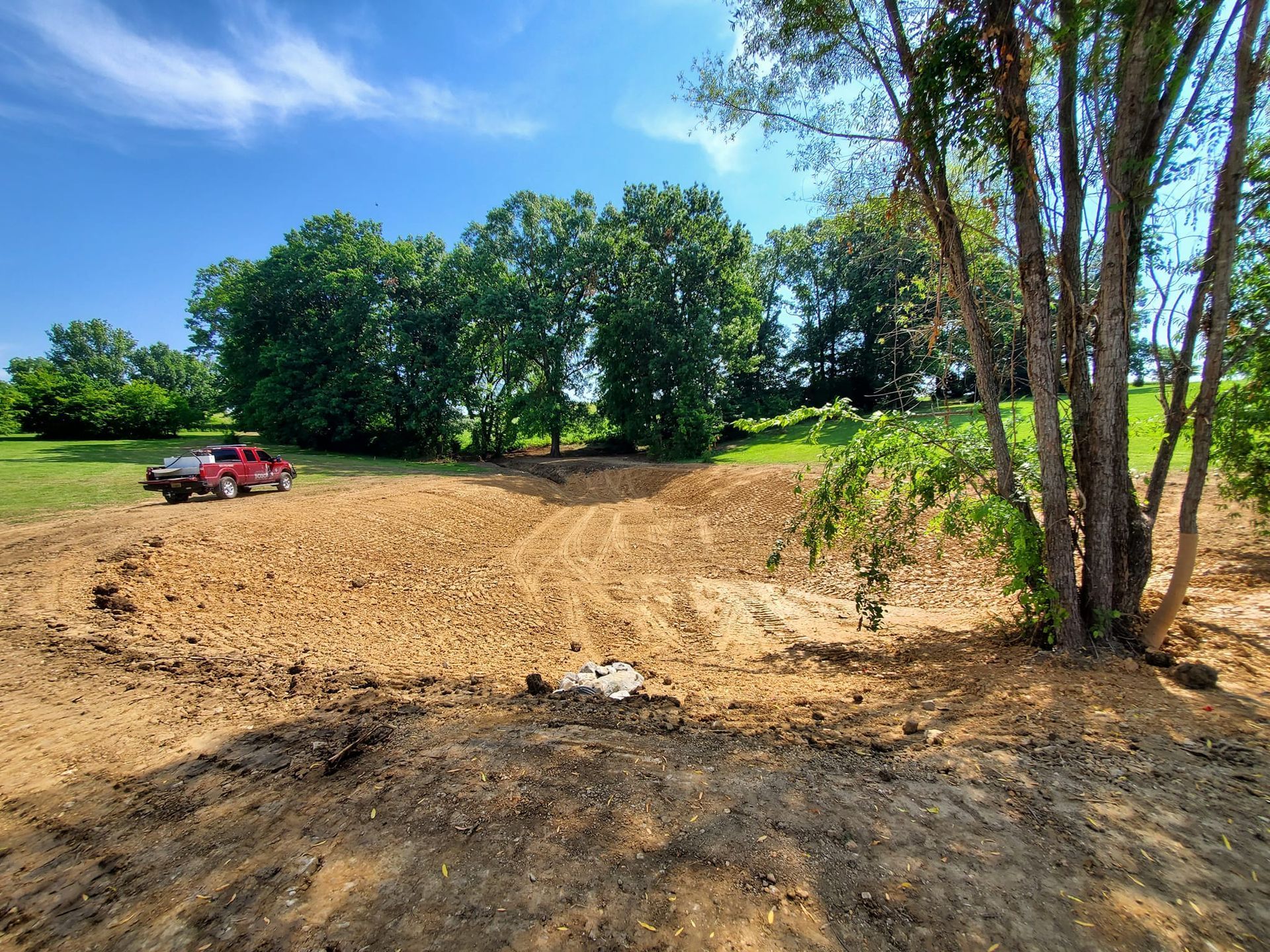 A red truck parked near a large, freshly excavated dirt clearing surrounded by trees under a bright blue sky.