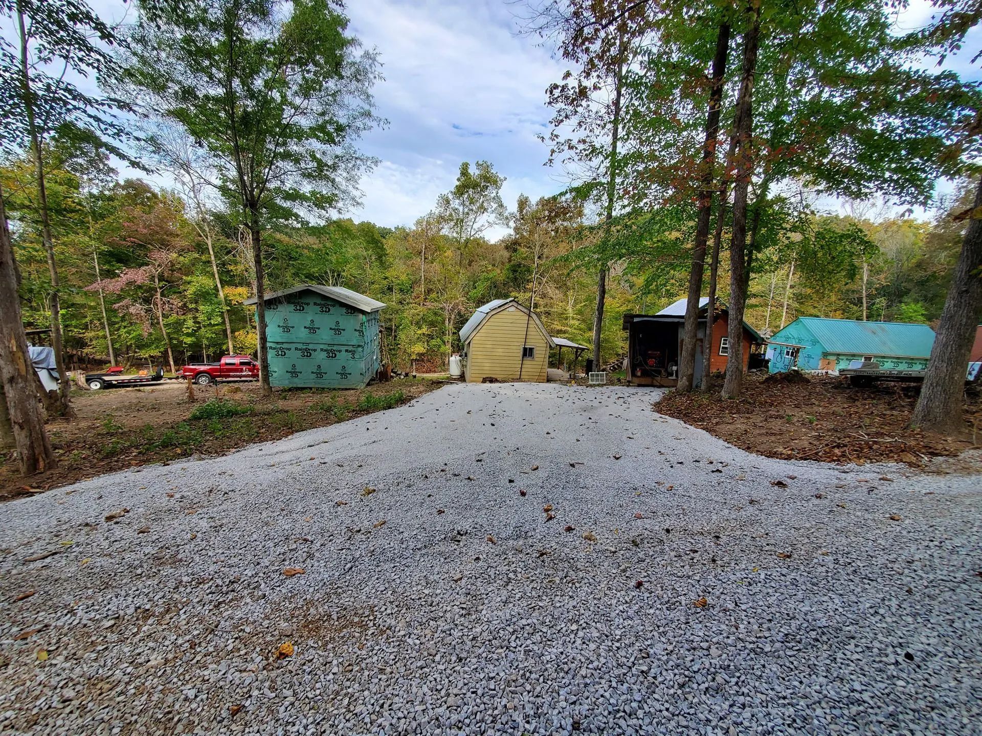 A gravel driveway leads through a wooded area toward three small sheds and a parked red vehicle under an autumn sky.
