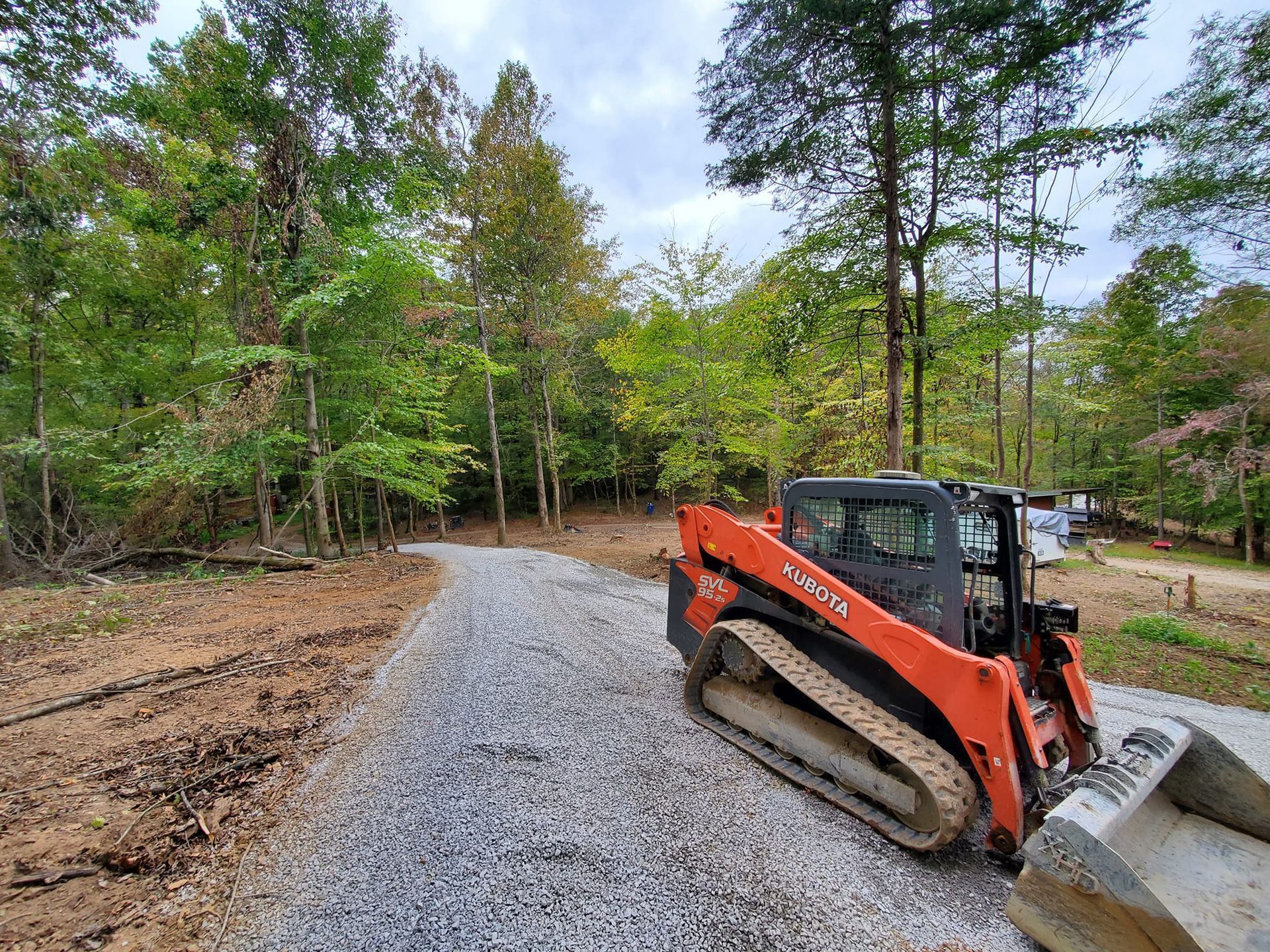 An orange tracked skid steer parked on a gravel path winding through a wooded area.