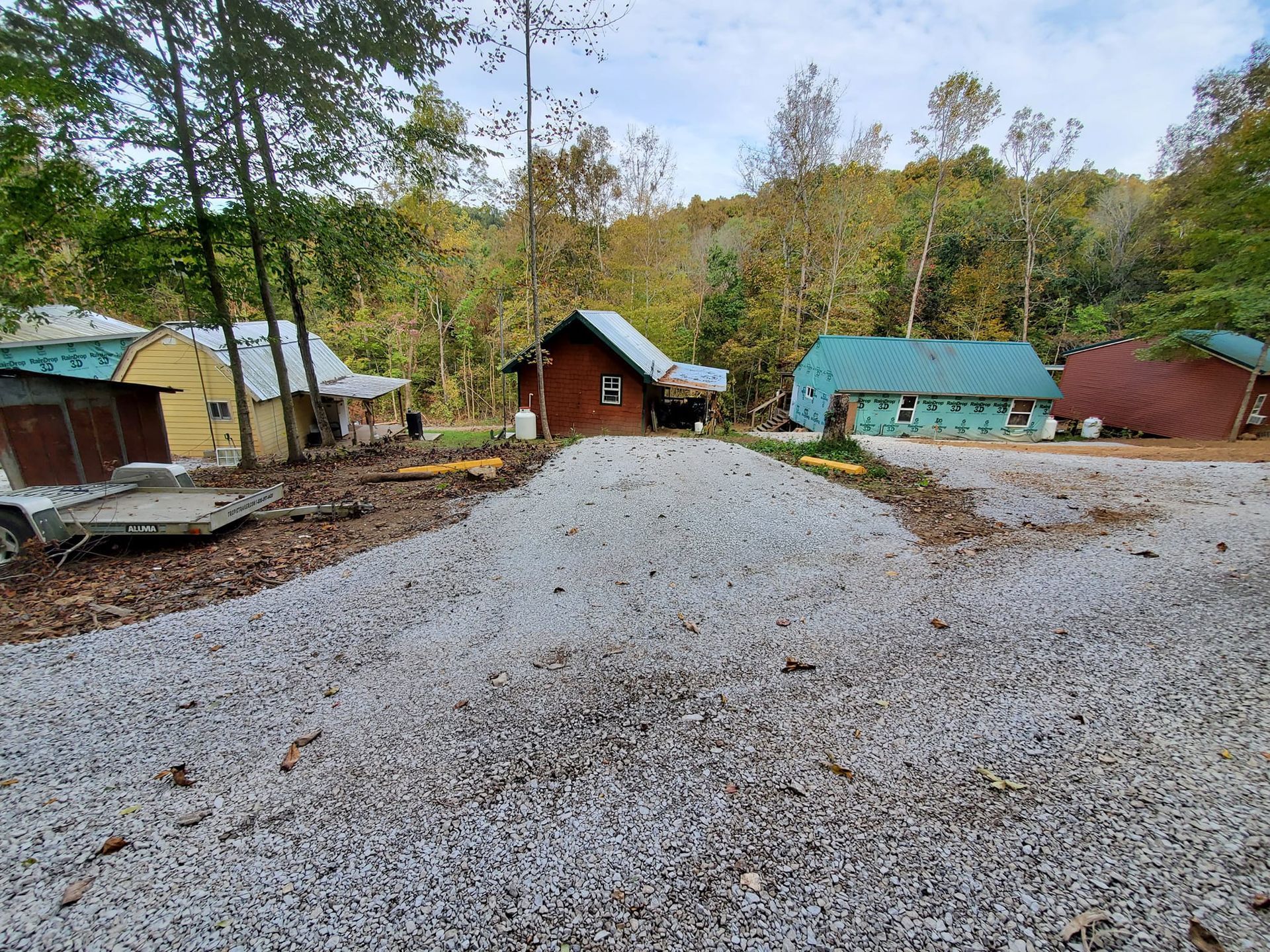 A gravel driveway leads toward a small brown cabin and a light blue shed, with trees and a red building in the background.