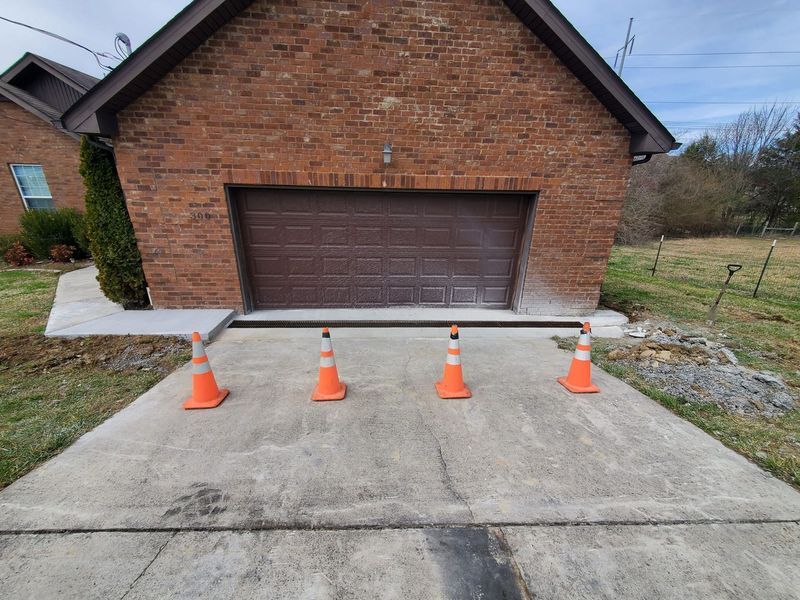A brick garage with a closed door, a new concrete trench drain, and four orange traffic cones on the driveway.