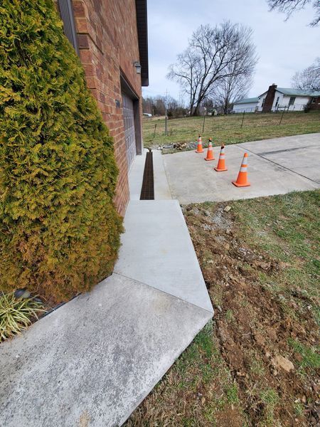 A concrete driveway with a metal trench drain in front of a brick garage, lined with traffic cones on the right.