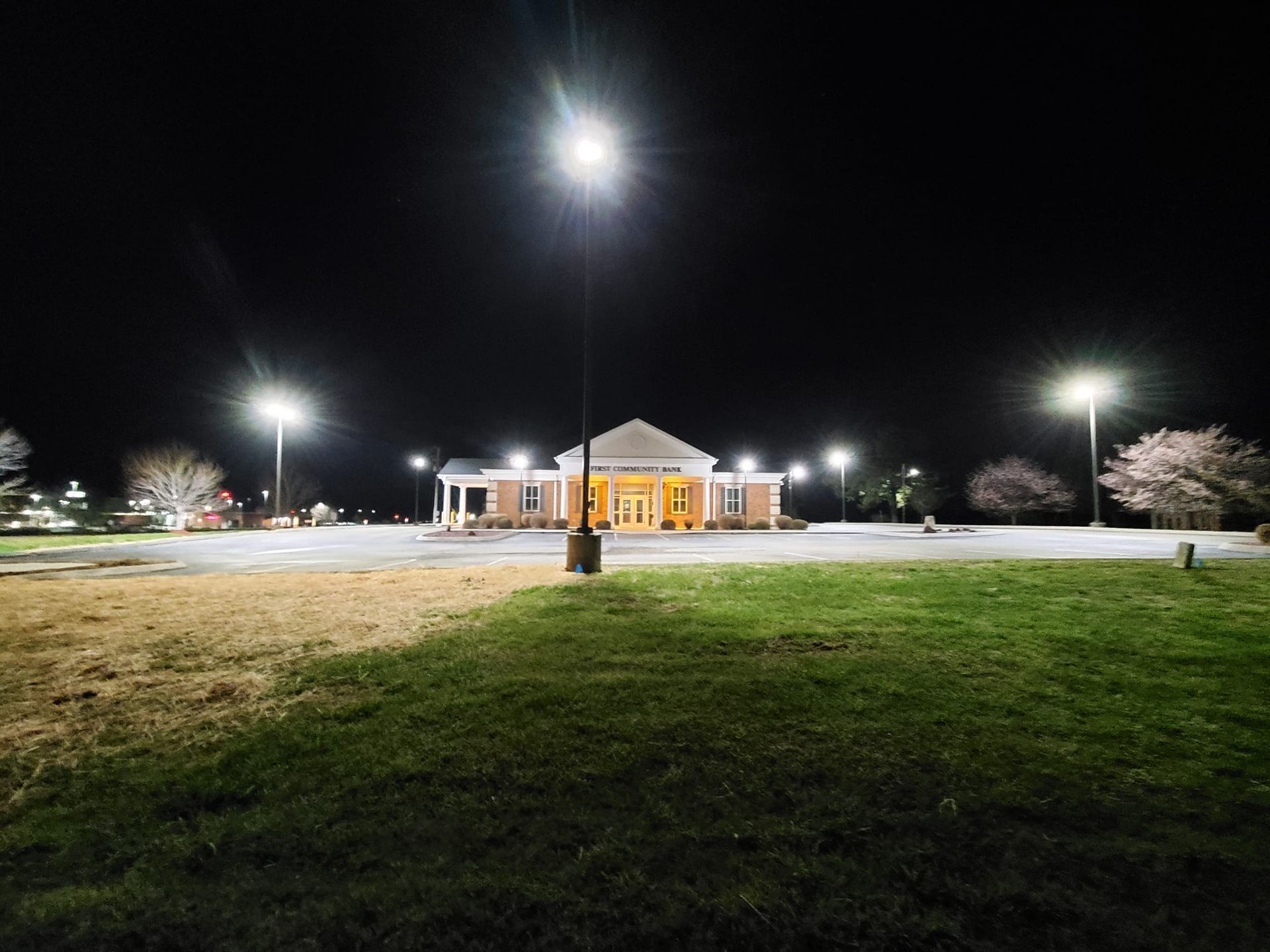 A brightly lit building stands at night, surrounded by a dark parking lot and grass, illuminated by tall streetlights.