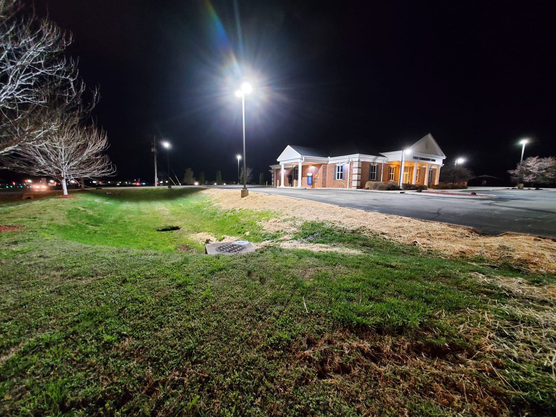 A parking lot and brick building illuminated by bright streetlights at night, viewed from a grassy foreground.