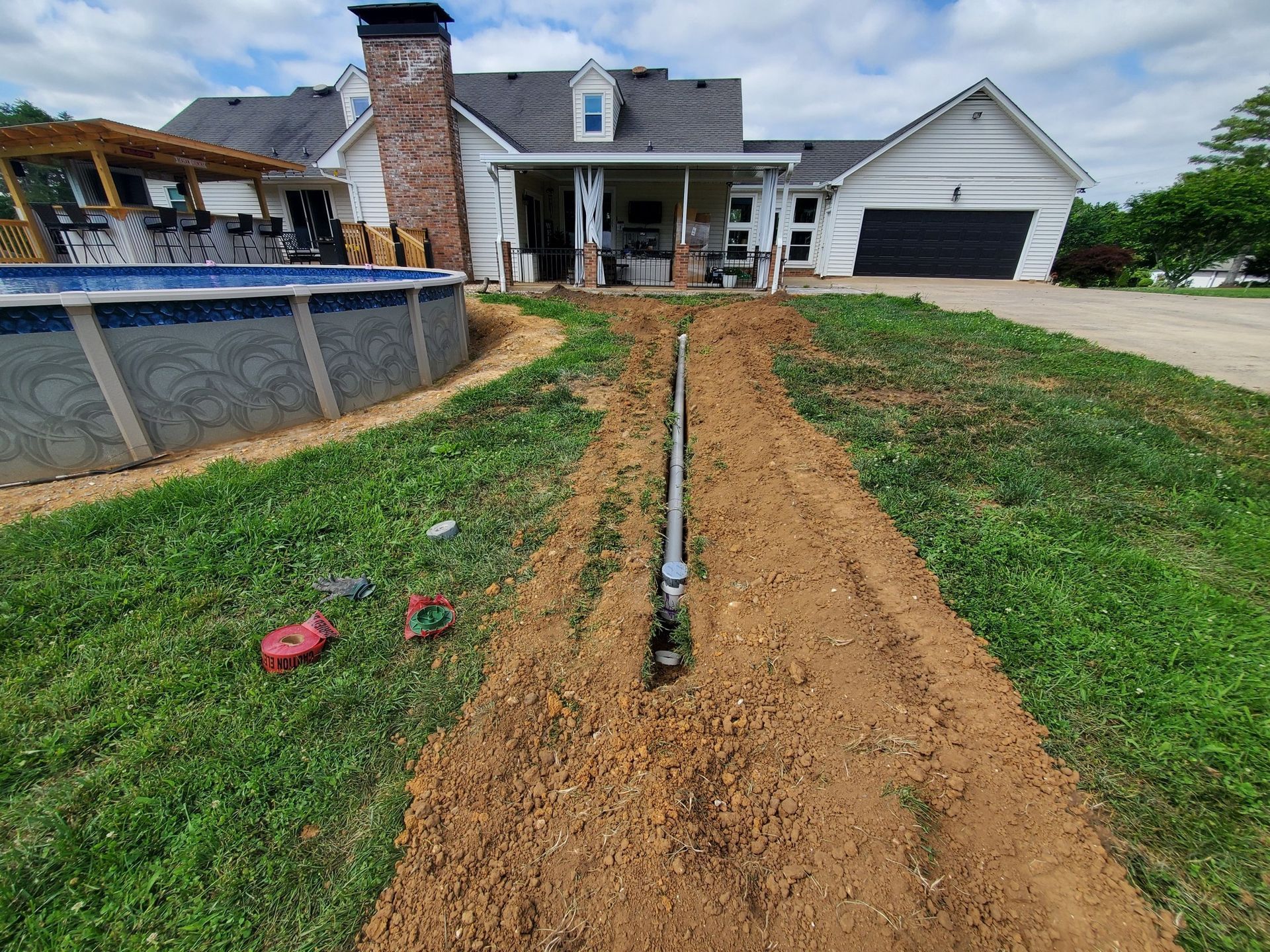 A trench with a pipe runs through a lawn toward a house with a pool and patio area.