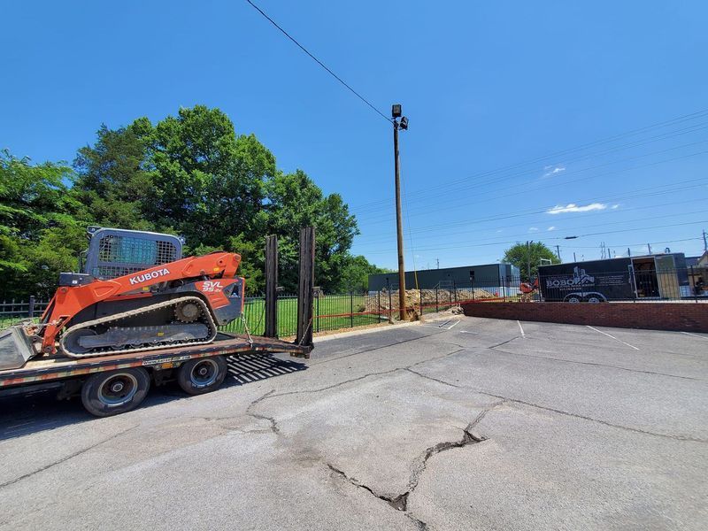 An orange skid-steer loader sits on a trailer in a sunny parking lot next to a utility pole and a construction site.