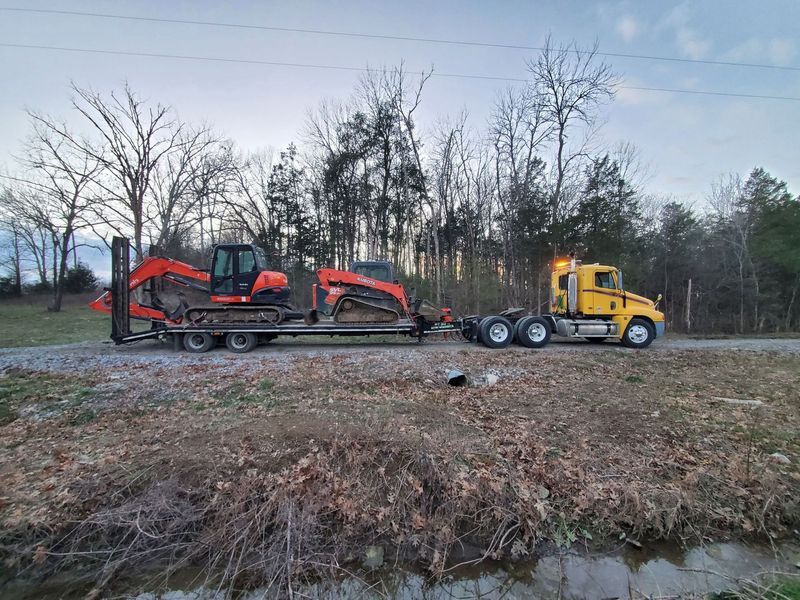 A yellow semi-truck hauls an orange excavator and a small tracked loader on a flatbed trailer along a rural dirt road.