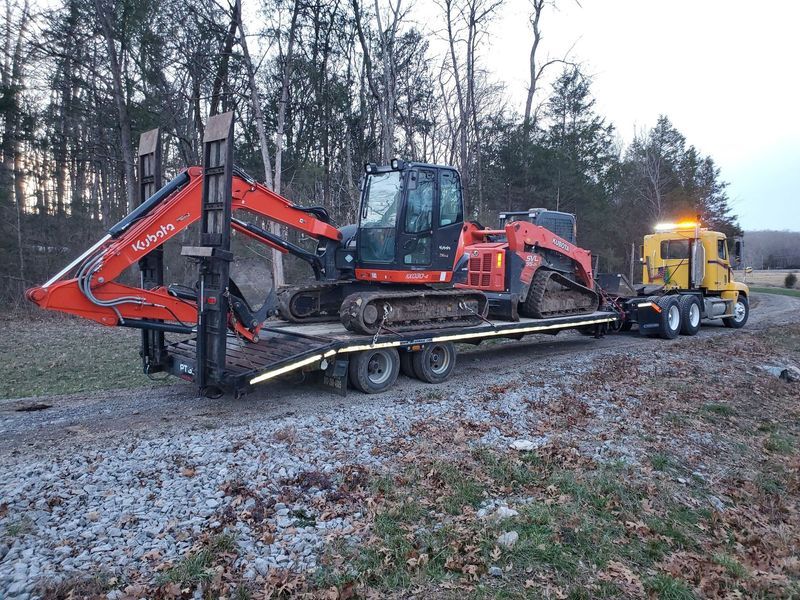 A bright orange Kubota excavator sits on a flatbed trailer being towed by a yellow truck on a gravel road.