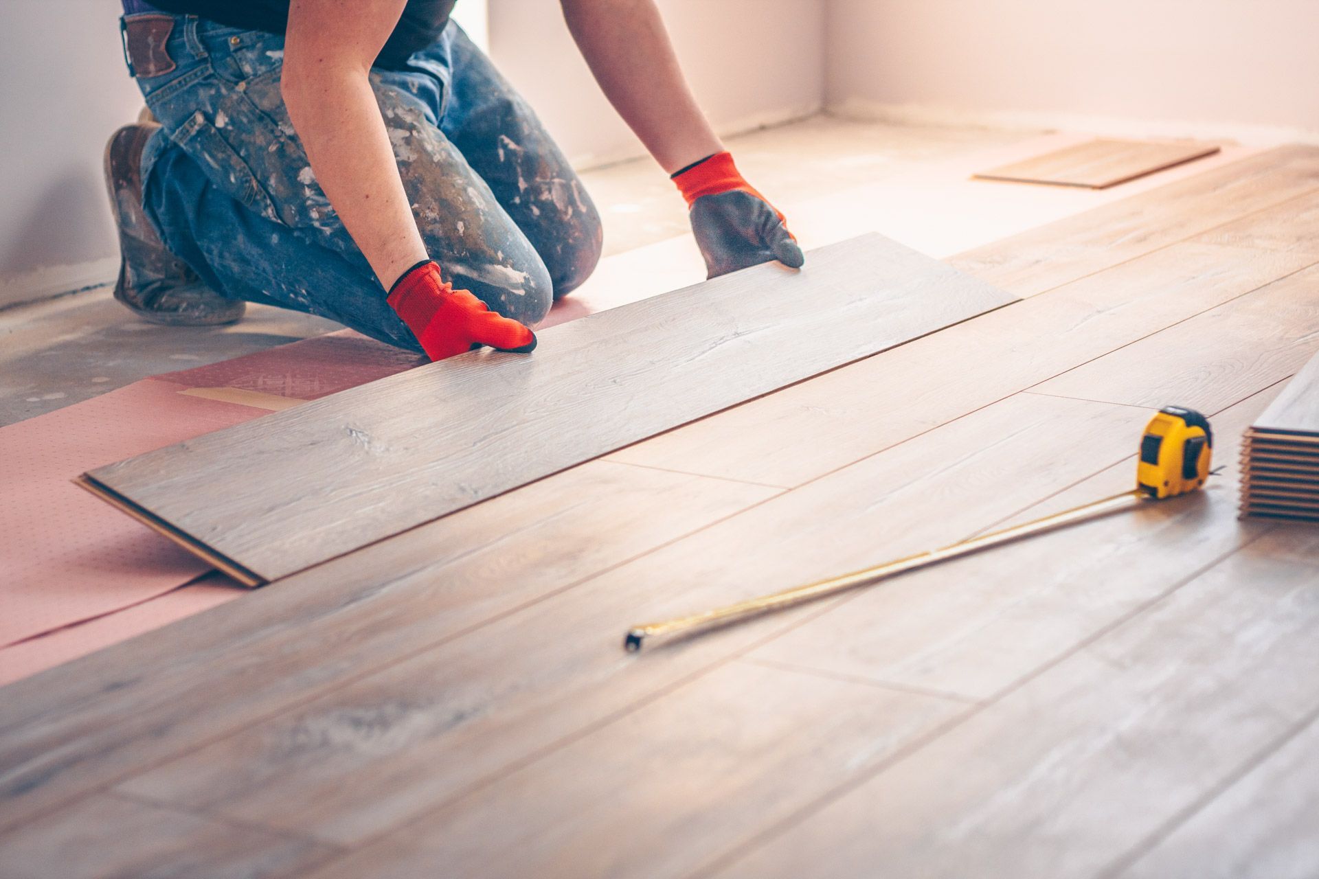 Person installing light wood-look flooring, kneeling with gloves, measuring tape nearby.