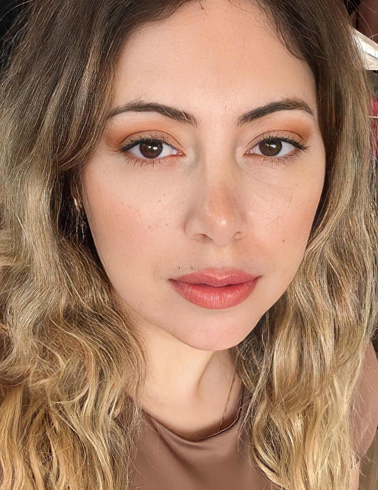 A close-up selfie of a person with long, wavy, light brown hair, wearing a neutral-toned top and subtle, warm makeup.