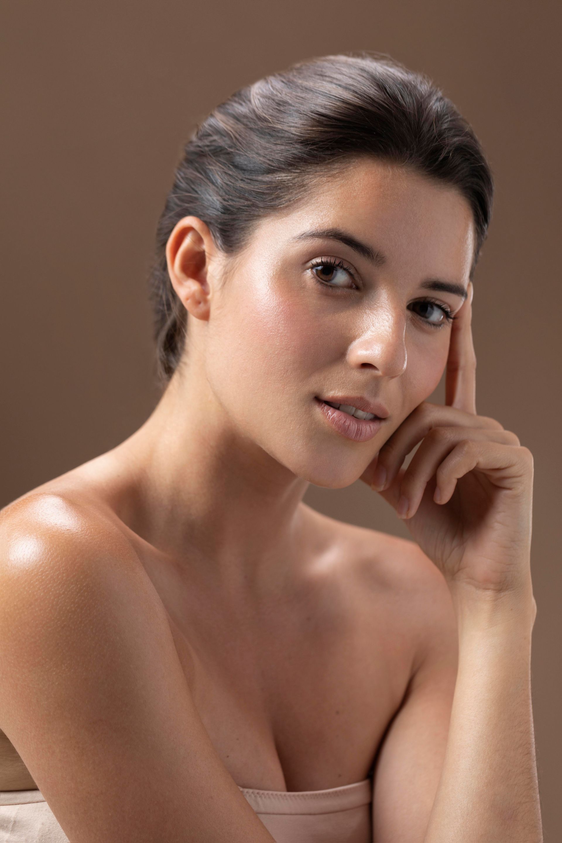 A person with dark hair pulled back poses against a brown background, resting their hand gently near their face.