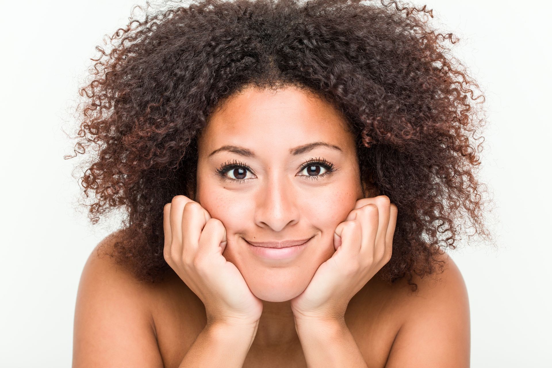 A person with curly hair smiling while resting their chin on both hands against a plain white background.