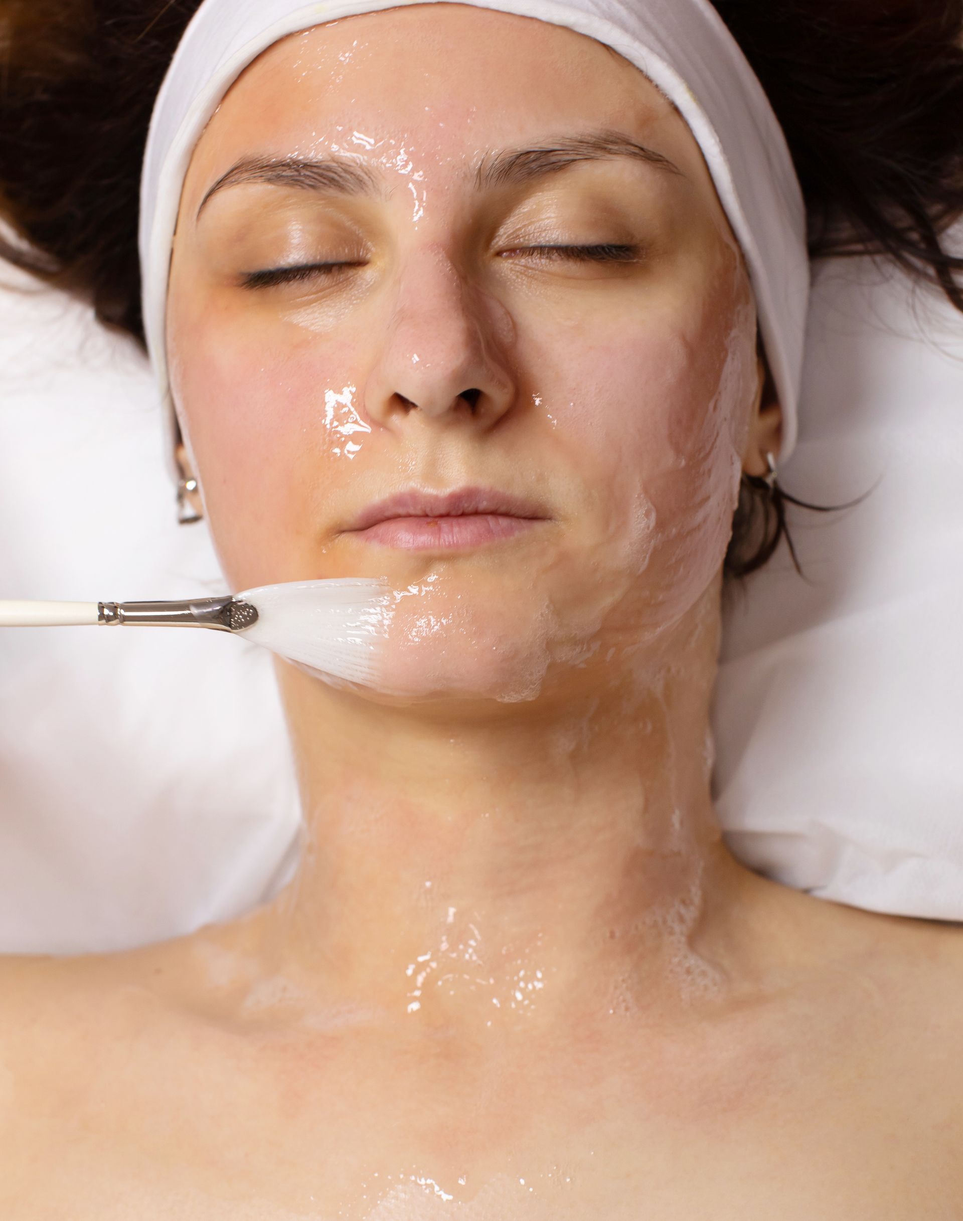 A person receiving a facial treatment as a brush applies a clear, gel-like substance to their chin.
