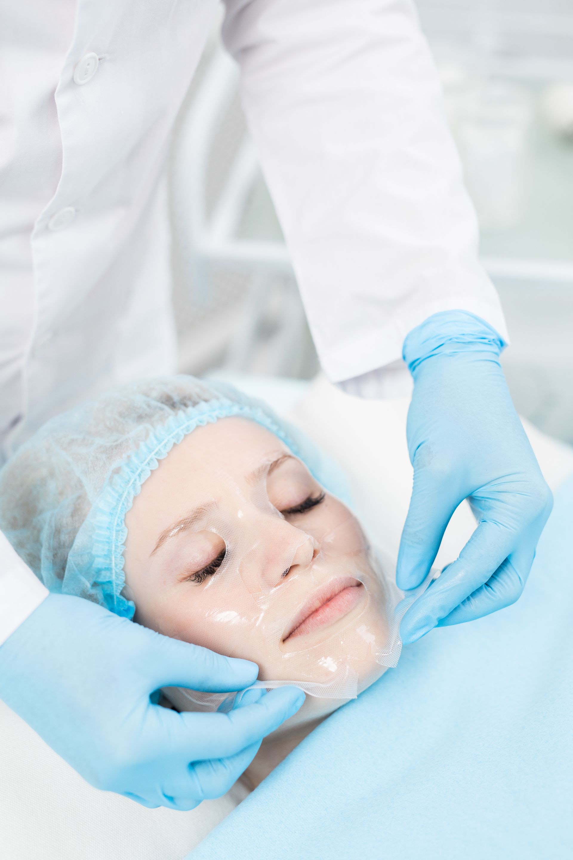A professional wearing blue gloves applies a clear sheet mask to a patient's face in a clinical setting.