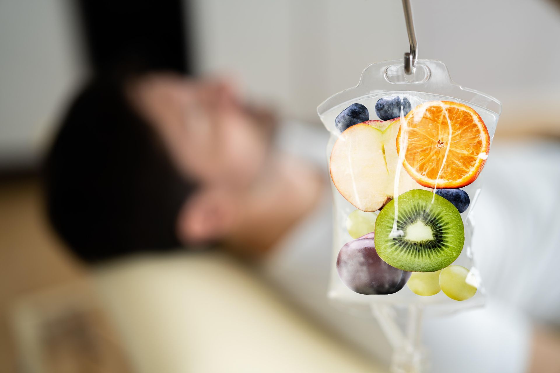A blurry person lies in the background while a clear IV bag containing various pieces of fresh fruit hangs in the front.