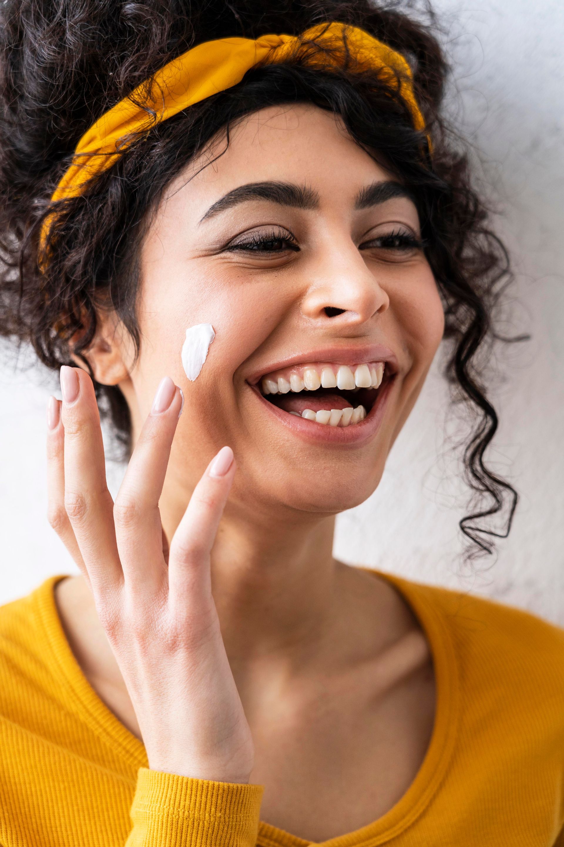 A smiling person wearing a yellow headband and top, applying a dab of white cream to their cheek.