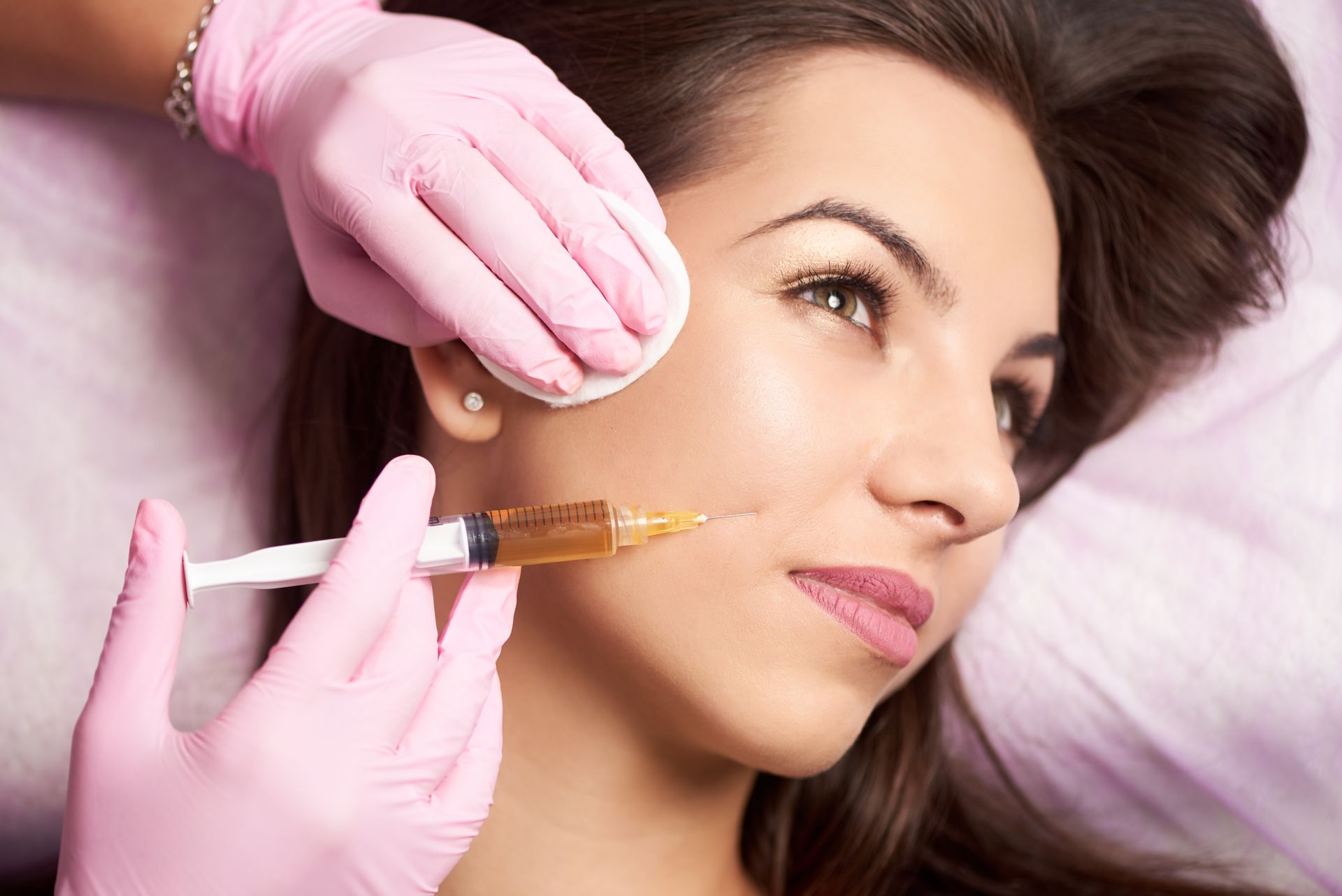 A person in pink gloves administers a facial injection while another hand holds a cotton pad to the recipient’s skin.