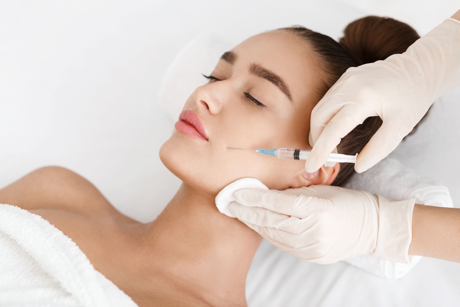 A professional in gloves administers a cosmetic facial injection to a person lying on a clinical treatment bed.