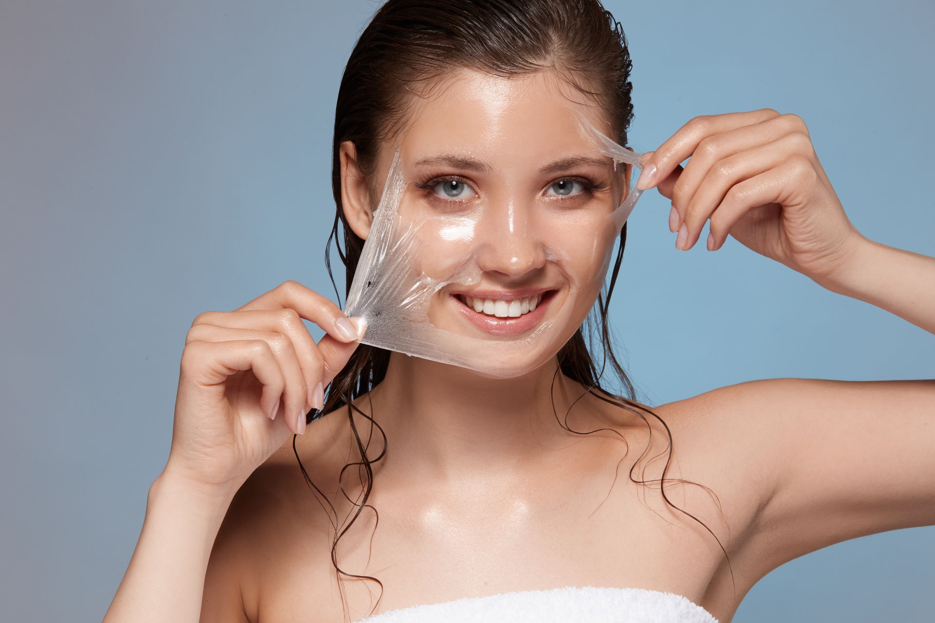 A person smiling while peeling a translucent, gel-like face mask off their skin against a blue background.
