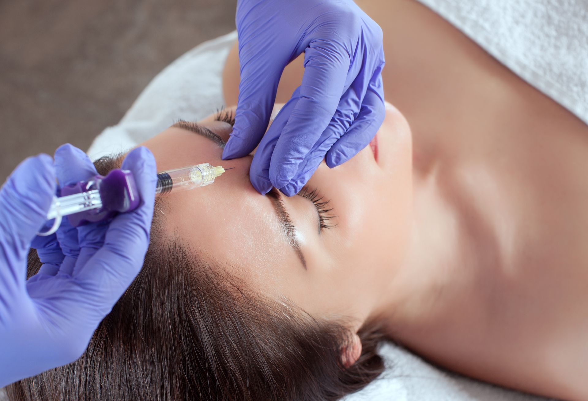 A medical professional in blue gloves administers a cosmetic injection to a patient’s forehead.
