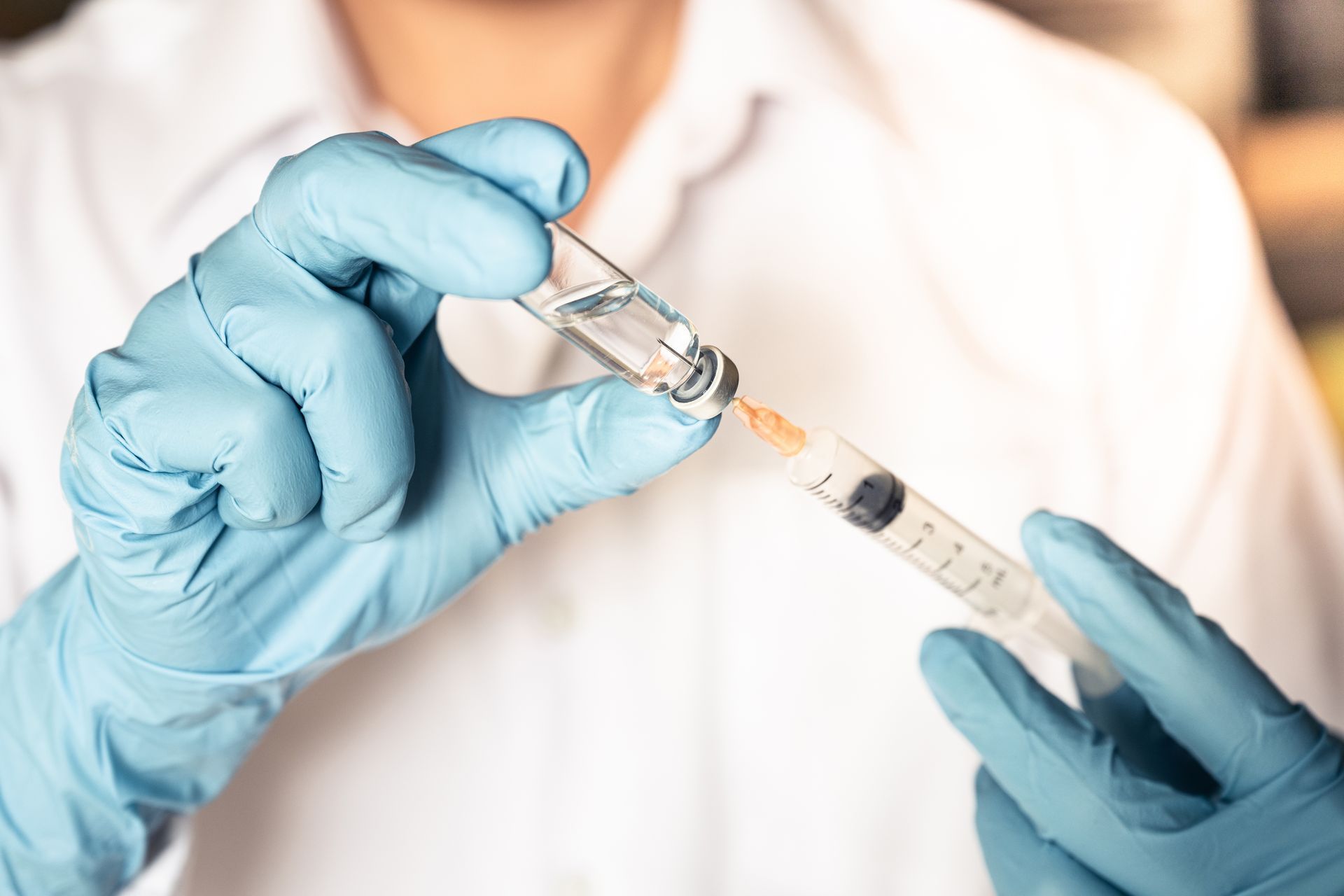 A person wearing blue medical gloves uses a syringe to draw liquid from a small glass vial.