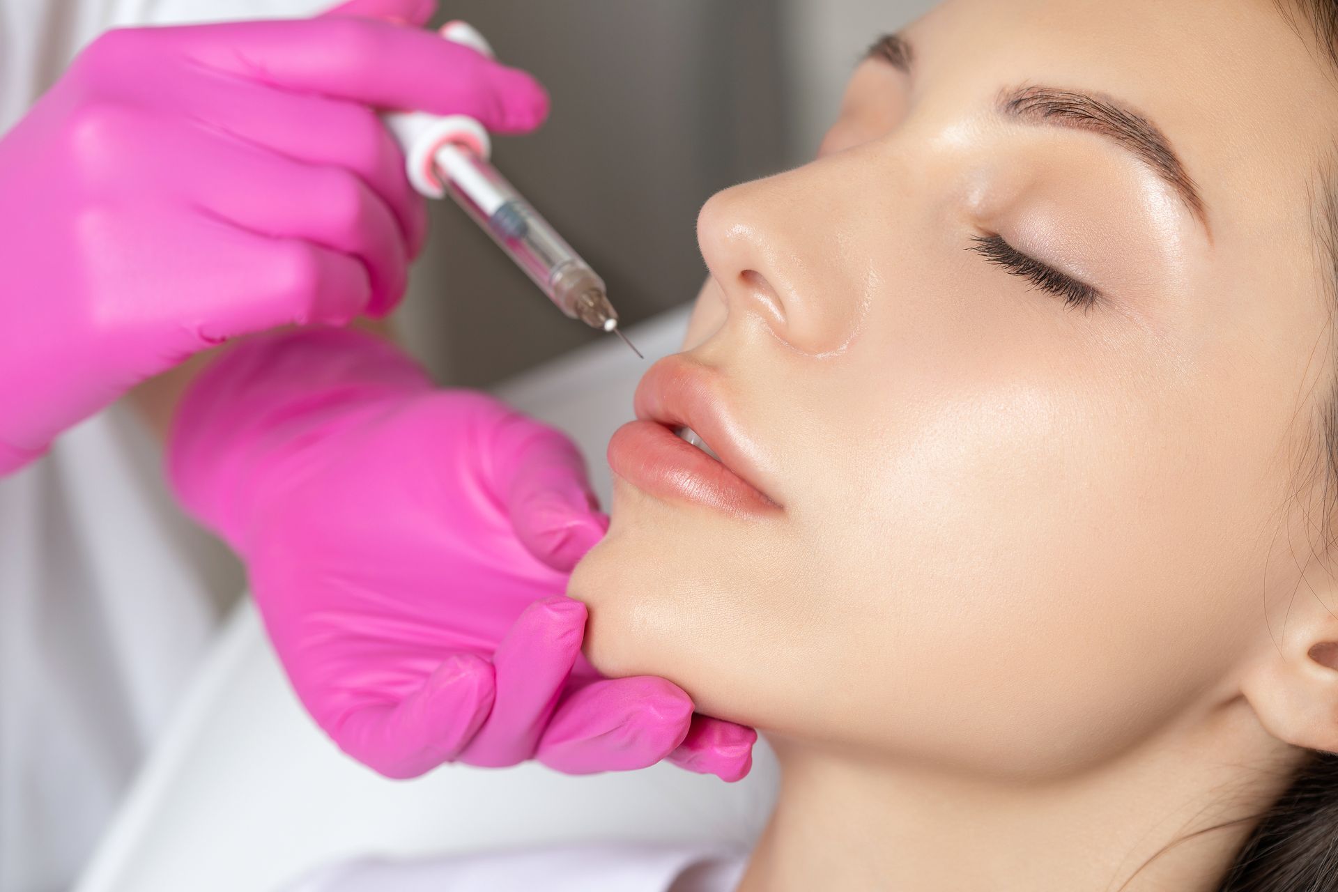 A medical professional in bright pink gloves administers a cosmetic filler injection into a person's upper lip.