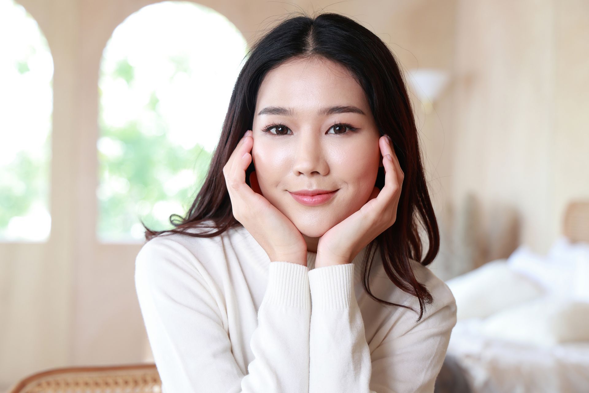 A person with long dark hair wears a white sweater, smiling while resting their chin on their hands in a bright room.