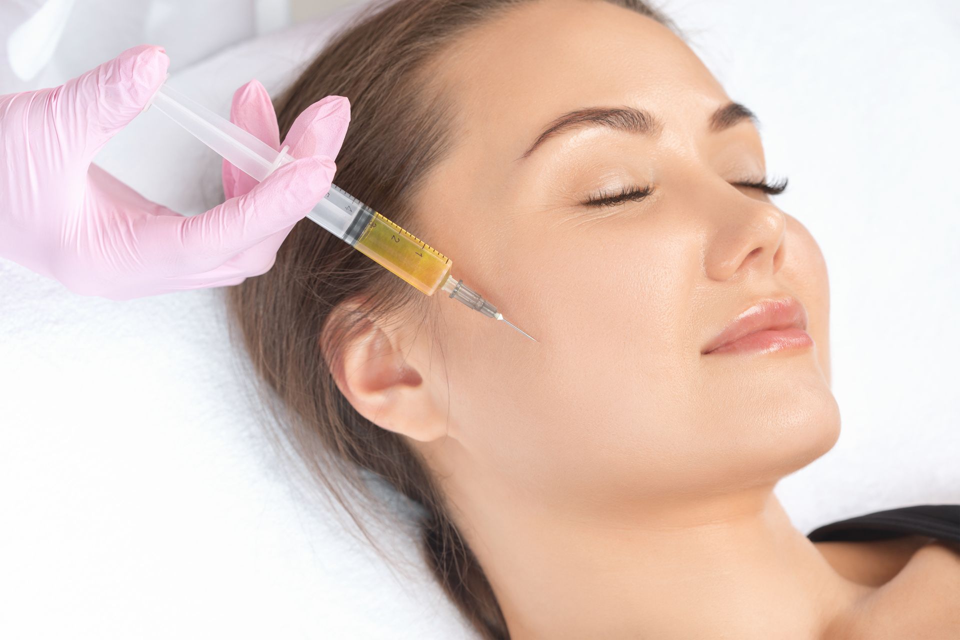 A person lying down receiving a facial injection from a gloved practitioner holding a syringe with yellowish fluid.