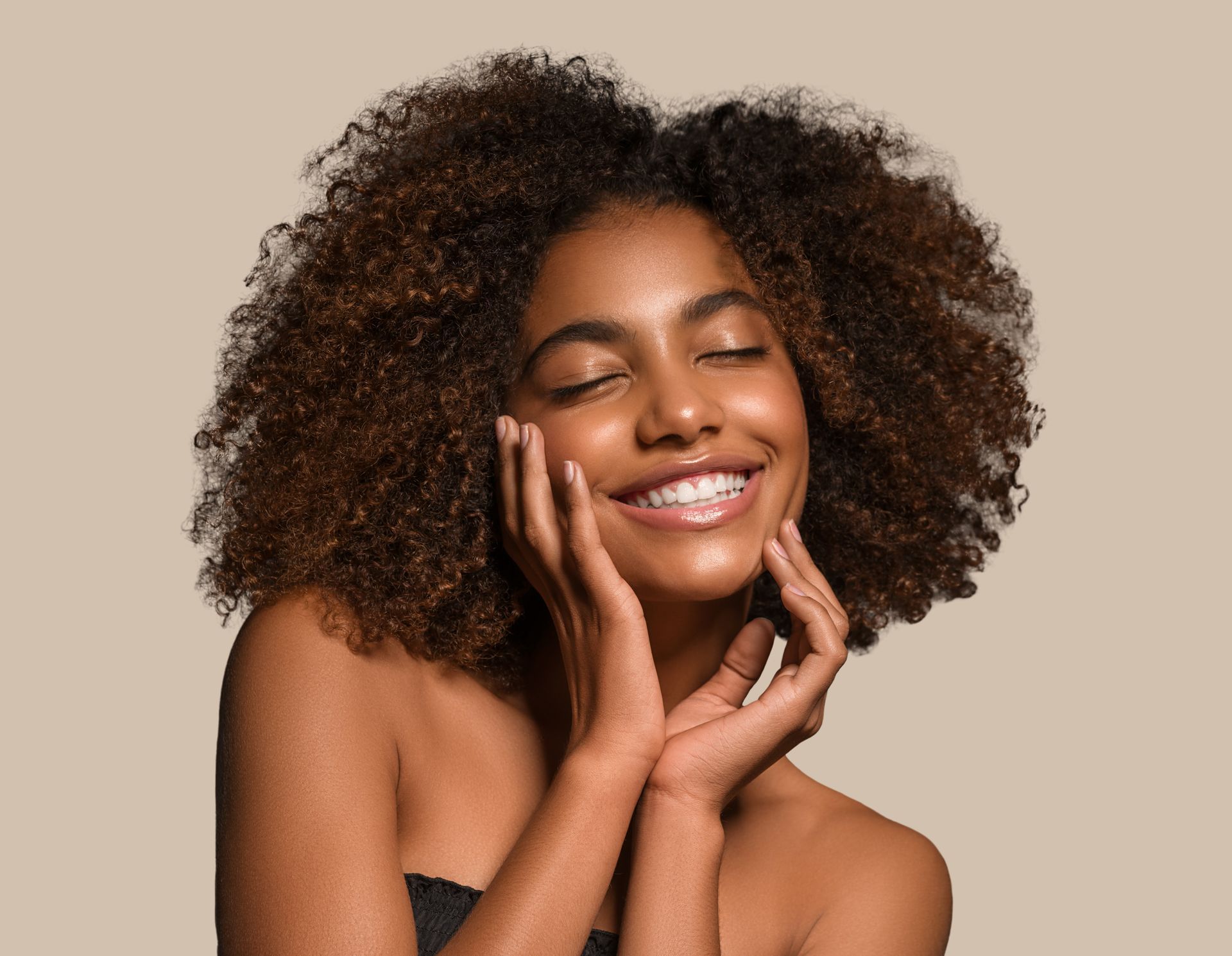 A smiling person with curly hair, hands cradling their face, against a solid beige background.