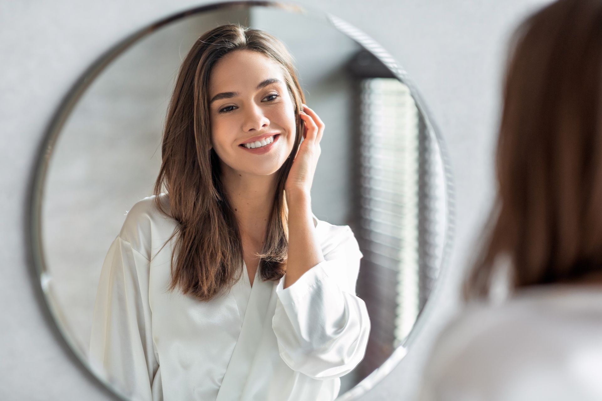 A person smiling while looking at their reflection in a circular mirror and touching their face.