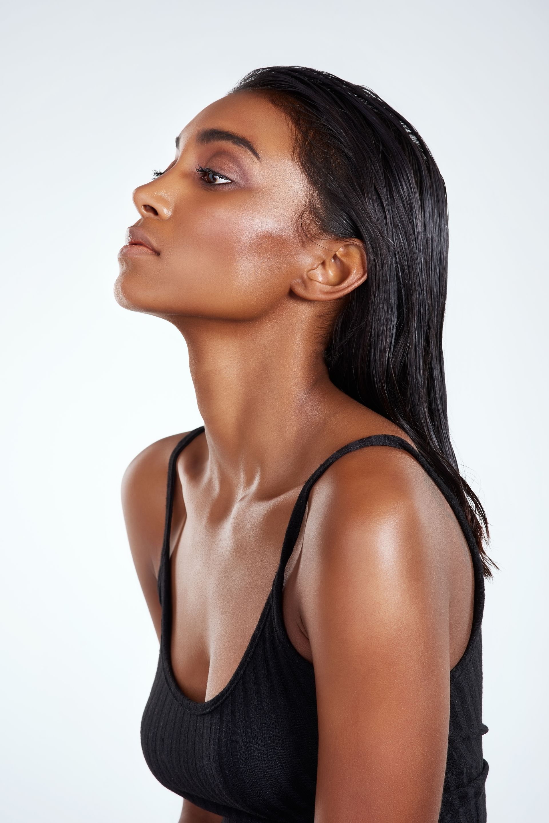 A person with wet, dark hair and glowing skin stands in profile against a light background, wearing a black tank top.