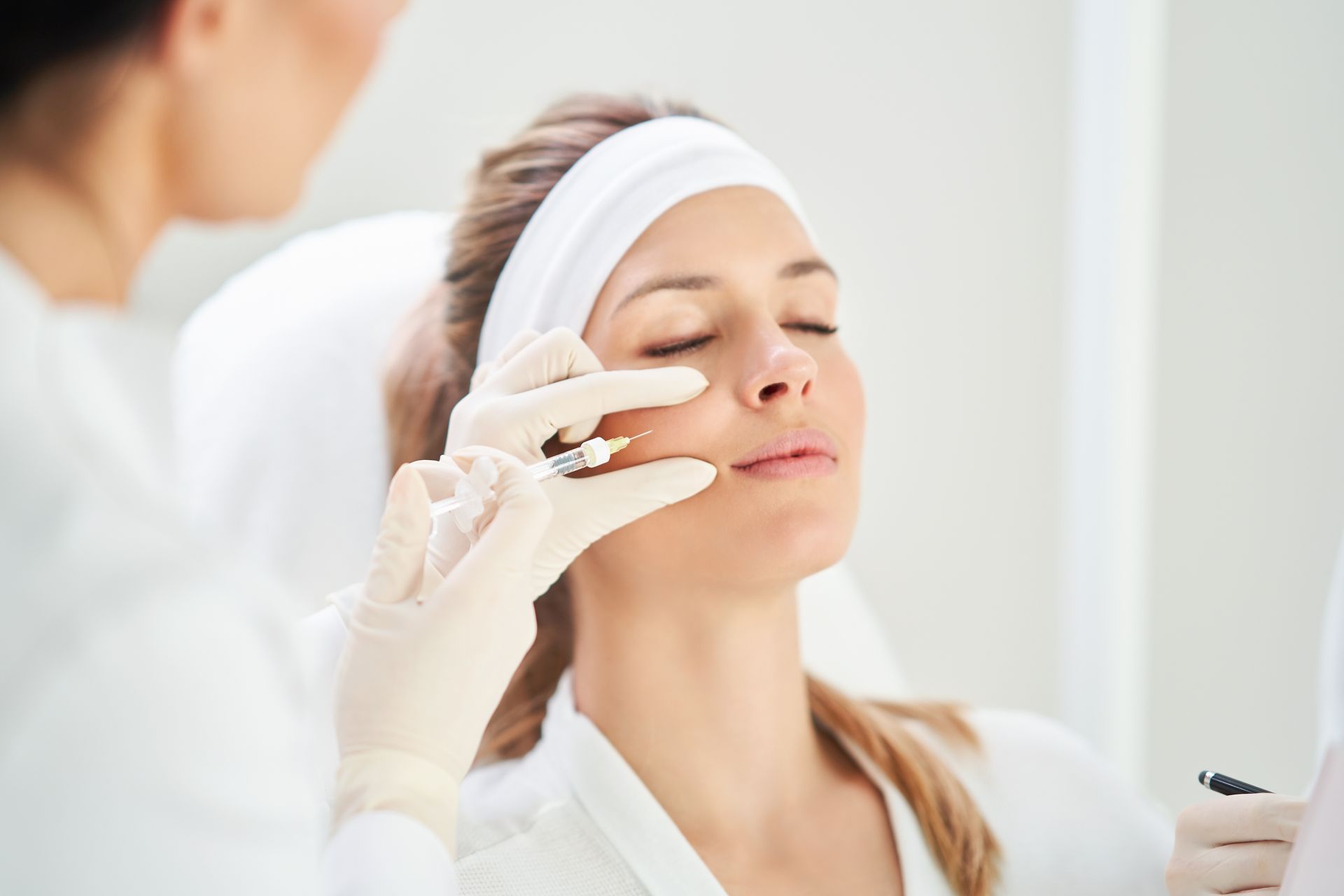 A medical professional in gloves administers a facial filler injection to a person wearing a white headband in a clinic.