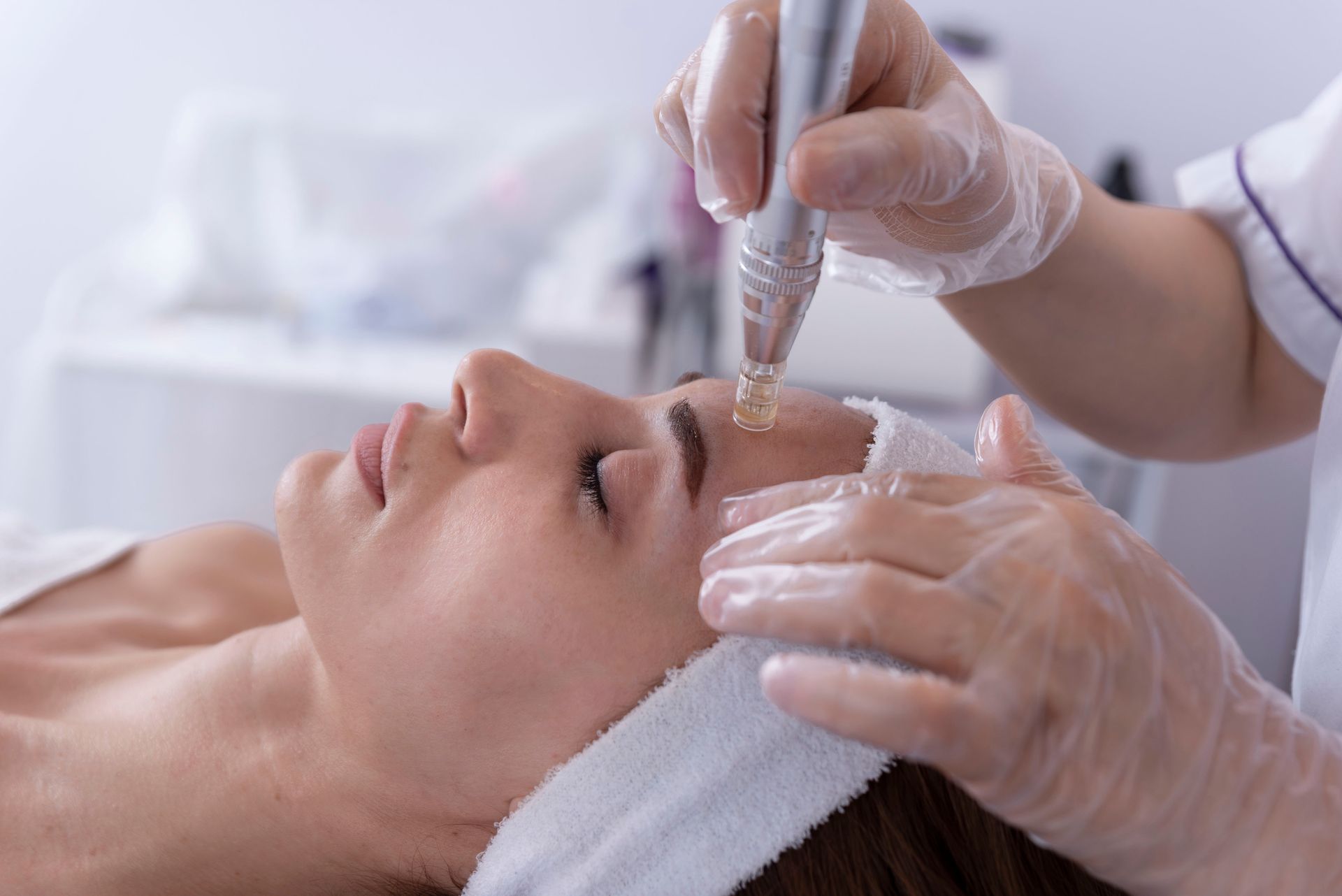 A professional in white gloves uses a handheld cosmetic device to apply product to a client’s forehead during a facial.