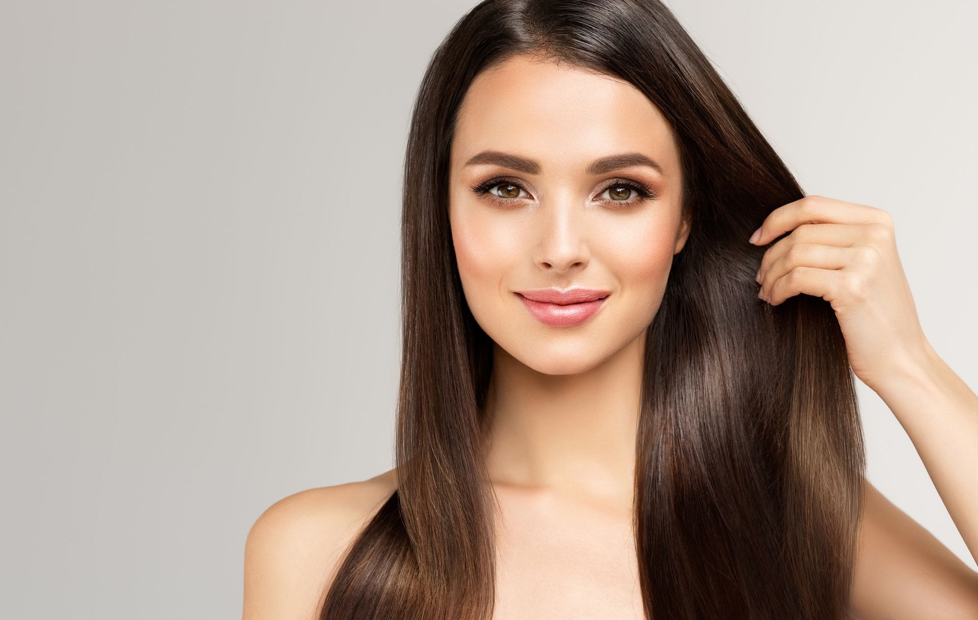 A person with long, straight dark brown hair smiles while holding a section of their hair against a light gray background.