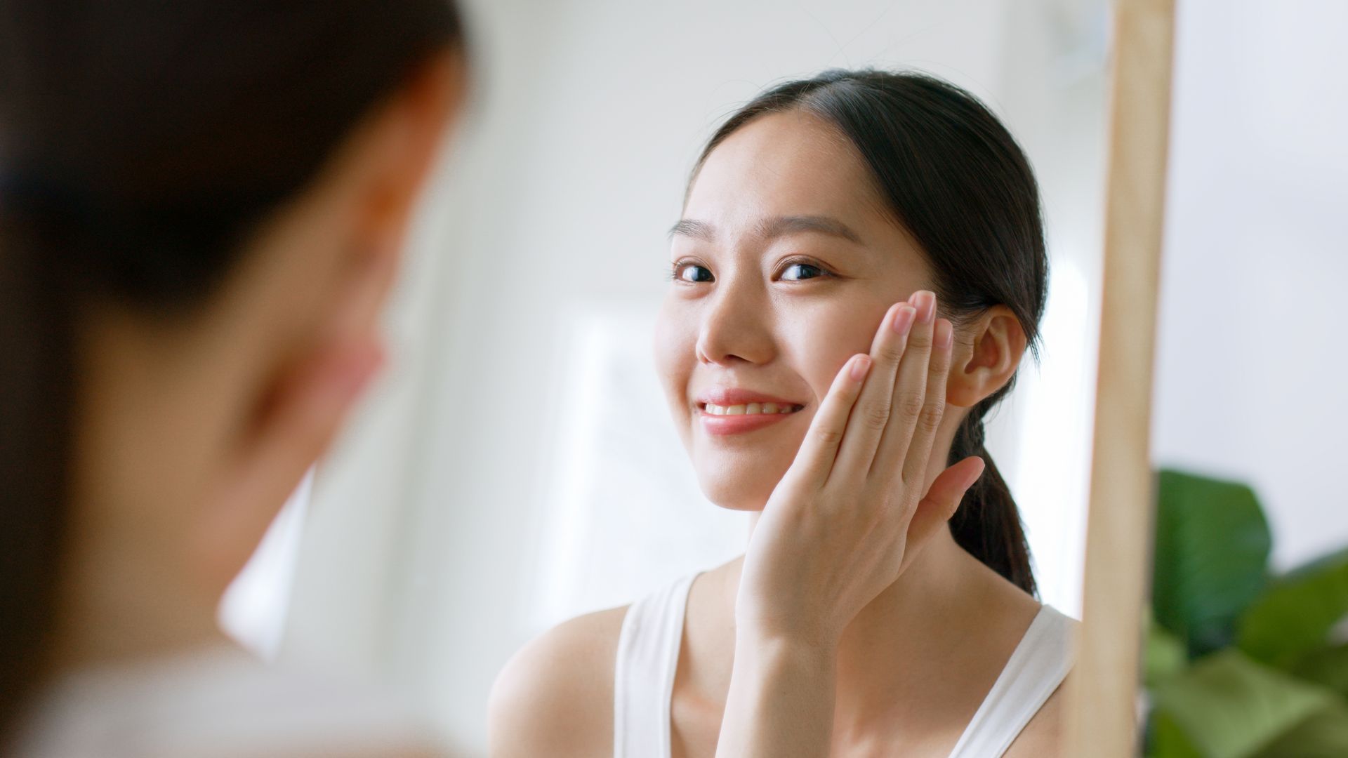 A person smiles while looking into a mirror, gently touching their cheek during a skincare routine.