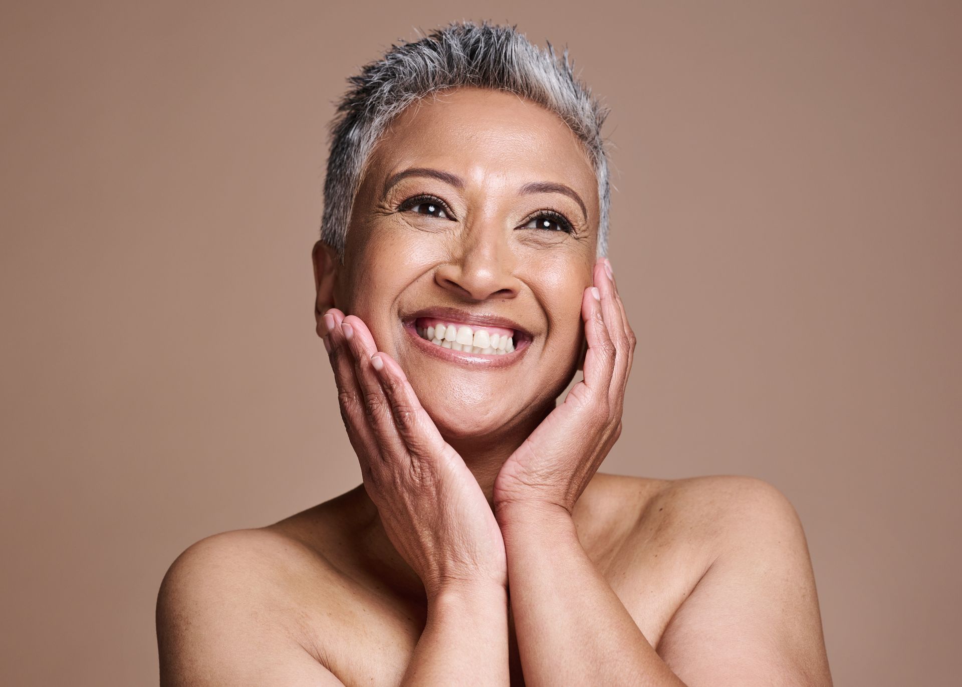 A person with short, grey hair smiling with their hands gently touching their cheeks against a solid neutral background.