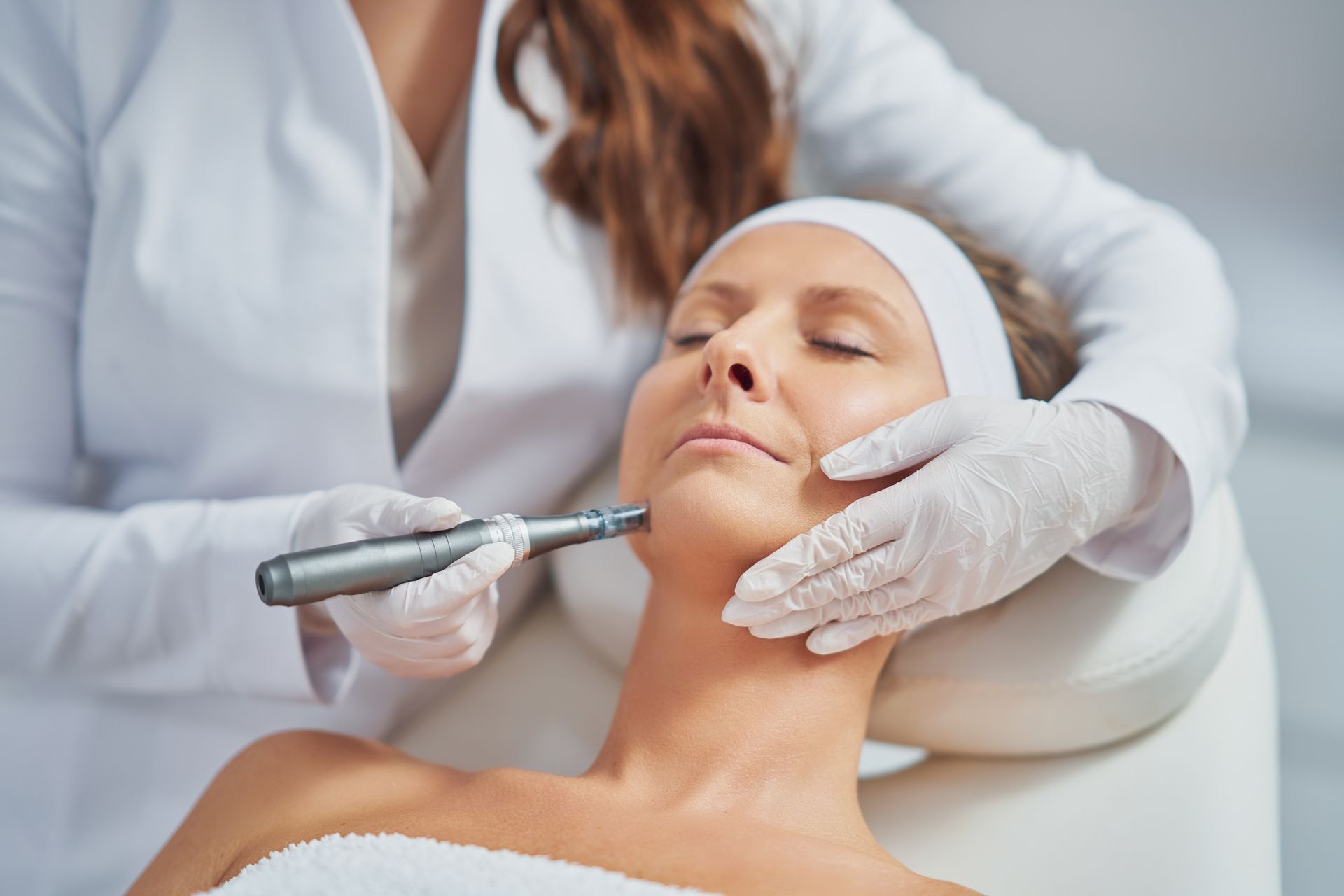 A professional aesthetician in white scrubs using a microneedling pen on a client's face in a clinical setting.