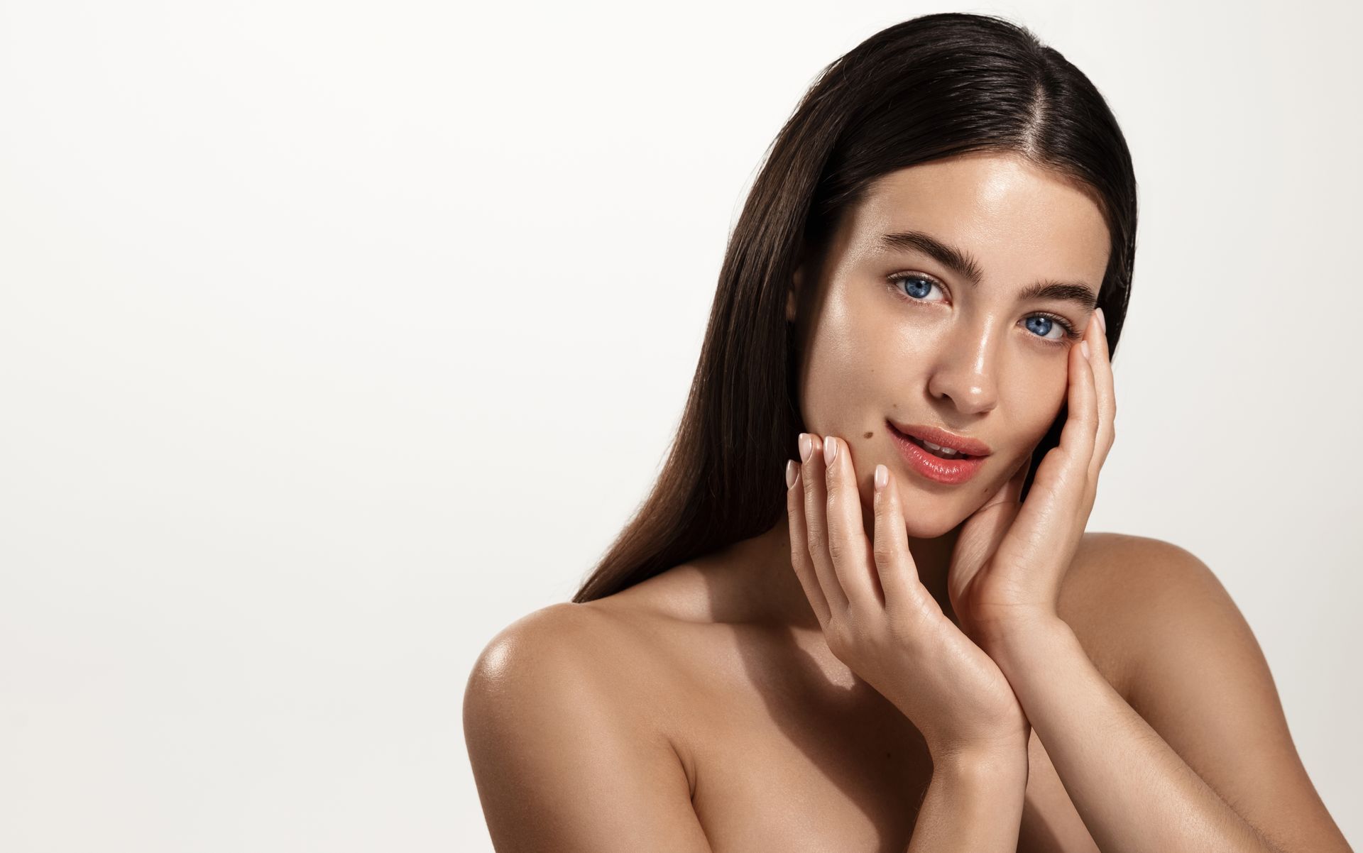 A person with dark hair and natural skin posing with hands gently touching their face against a plain light background.
