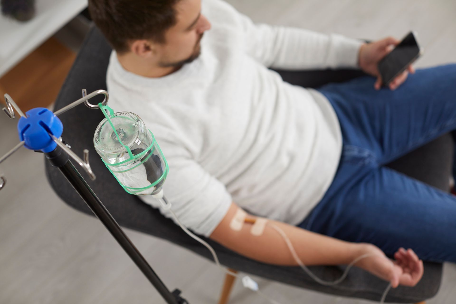 A person sitting in a chair receives an IV drip while holding a smartphone.