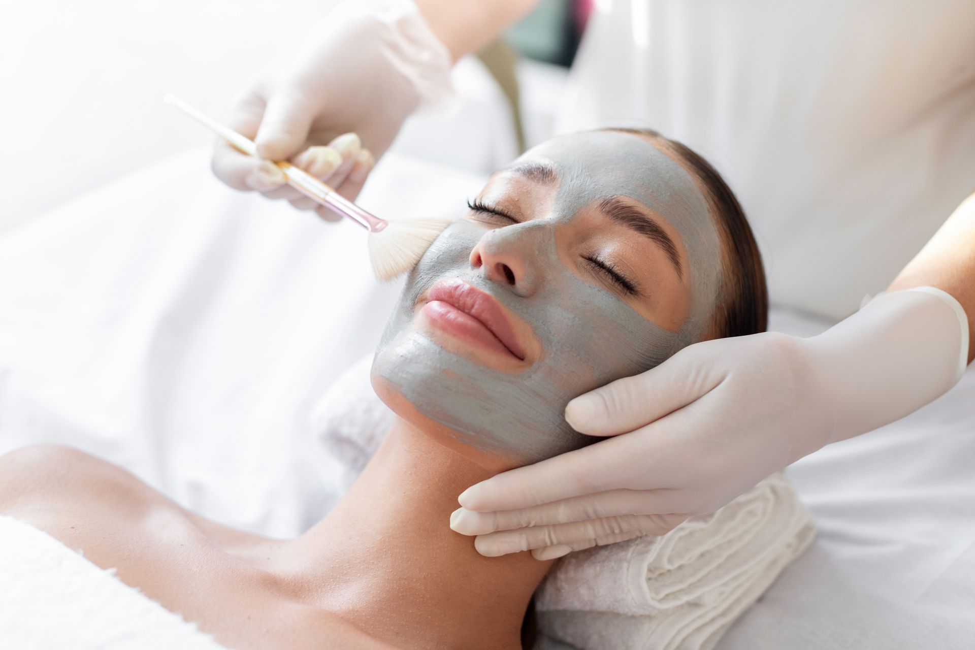 An esthetician in white gloves applies a gray clay face mask to a relaxed client using a cosmetic brush.