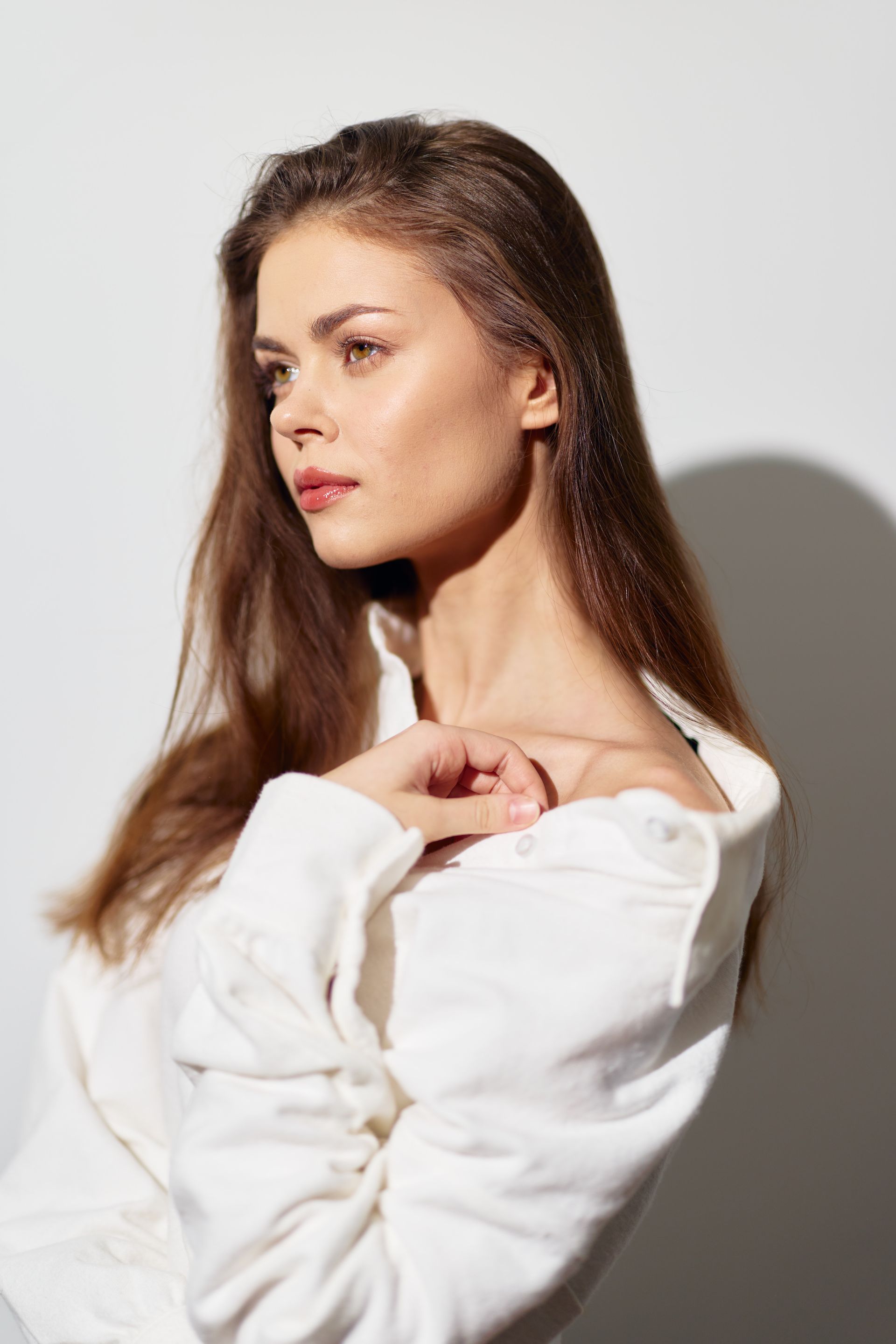 A person with long brown hair, wearing a white collared shirt, looks away against a neutral background.