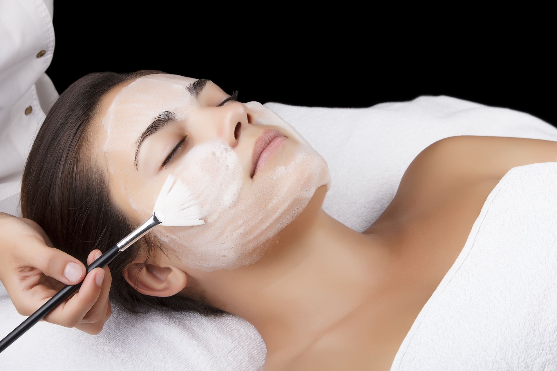 A spa professional uses a fan brush to apply a white facial mask onto a client's skin.