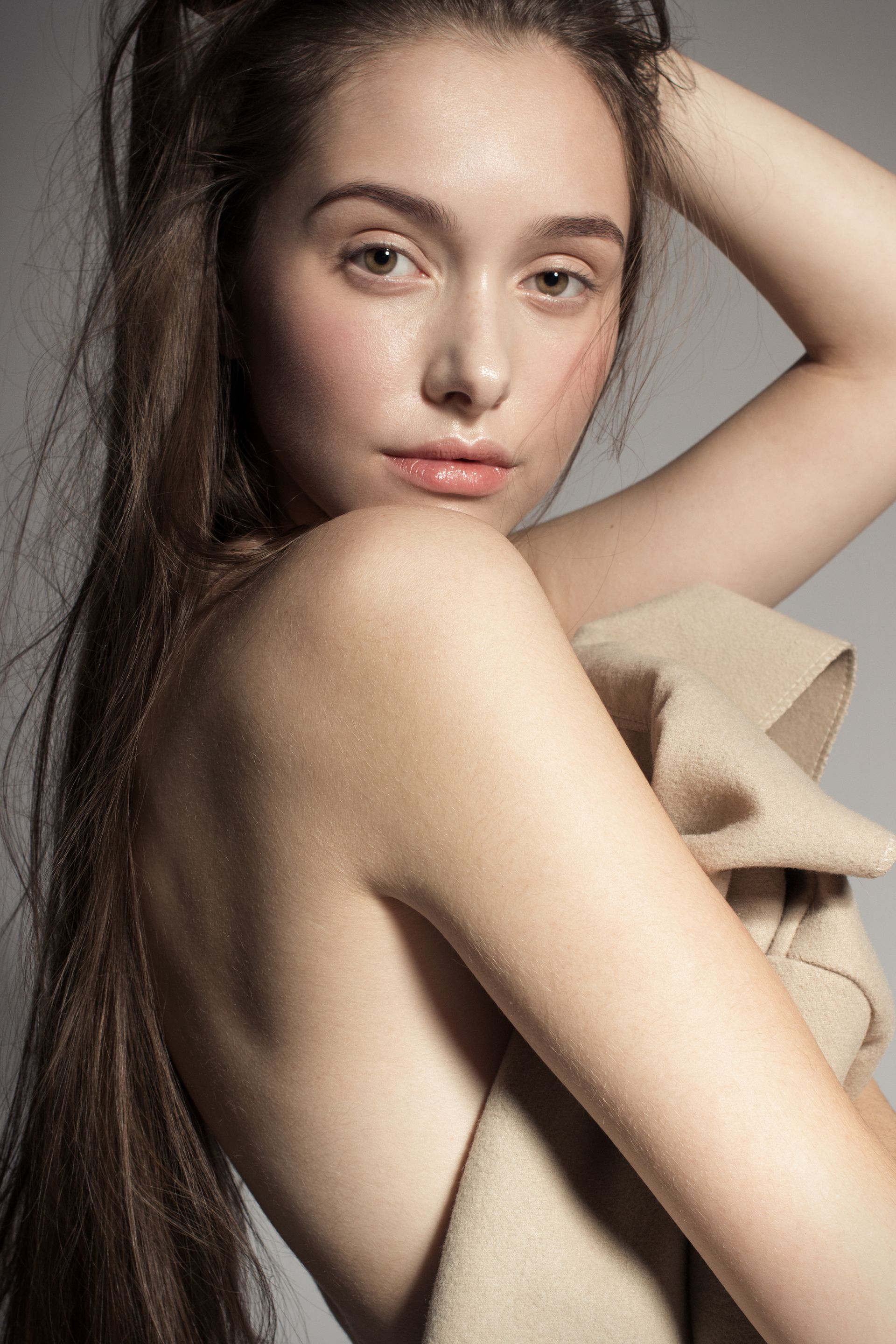 A person with long brown hair looks over their shoulder, draped in a tan fabric against a grey background.