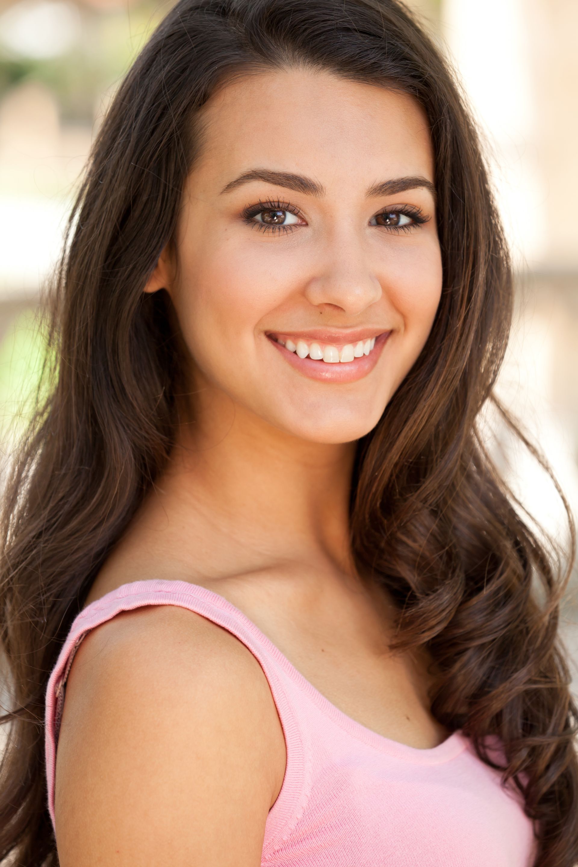 A smiling person with long, wavy dark brown hair wearing a pink top against a blurred background.
