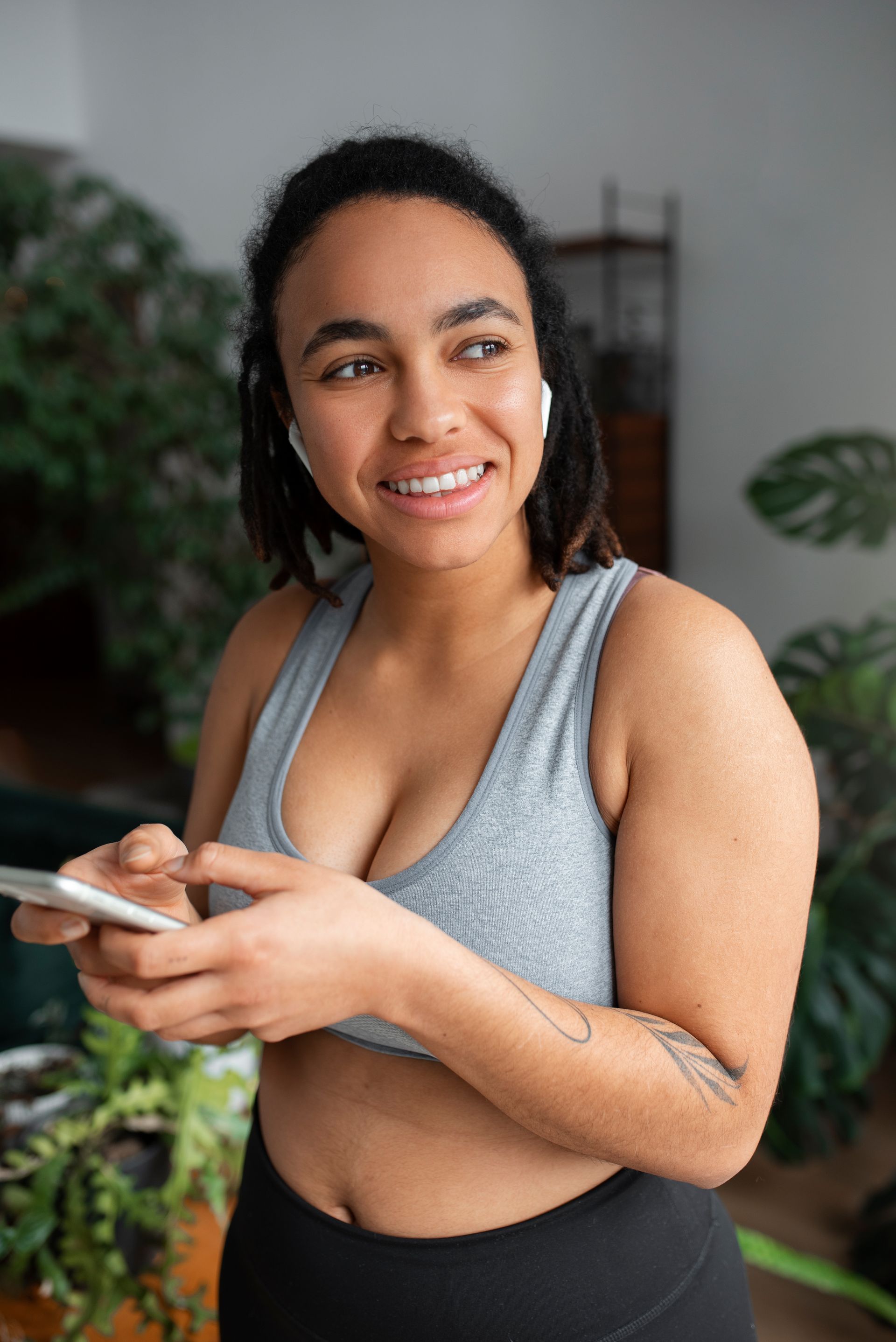 A person in a gray sports bra holds a smartphone, wearing wireless earbuds and smiling while looking to the side.