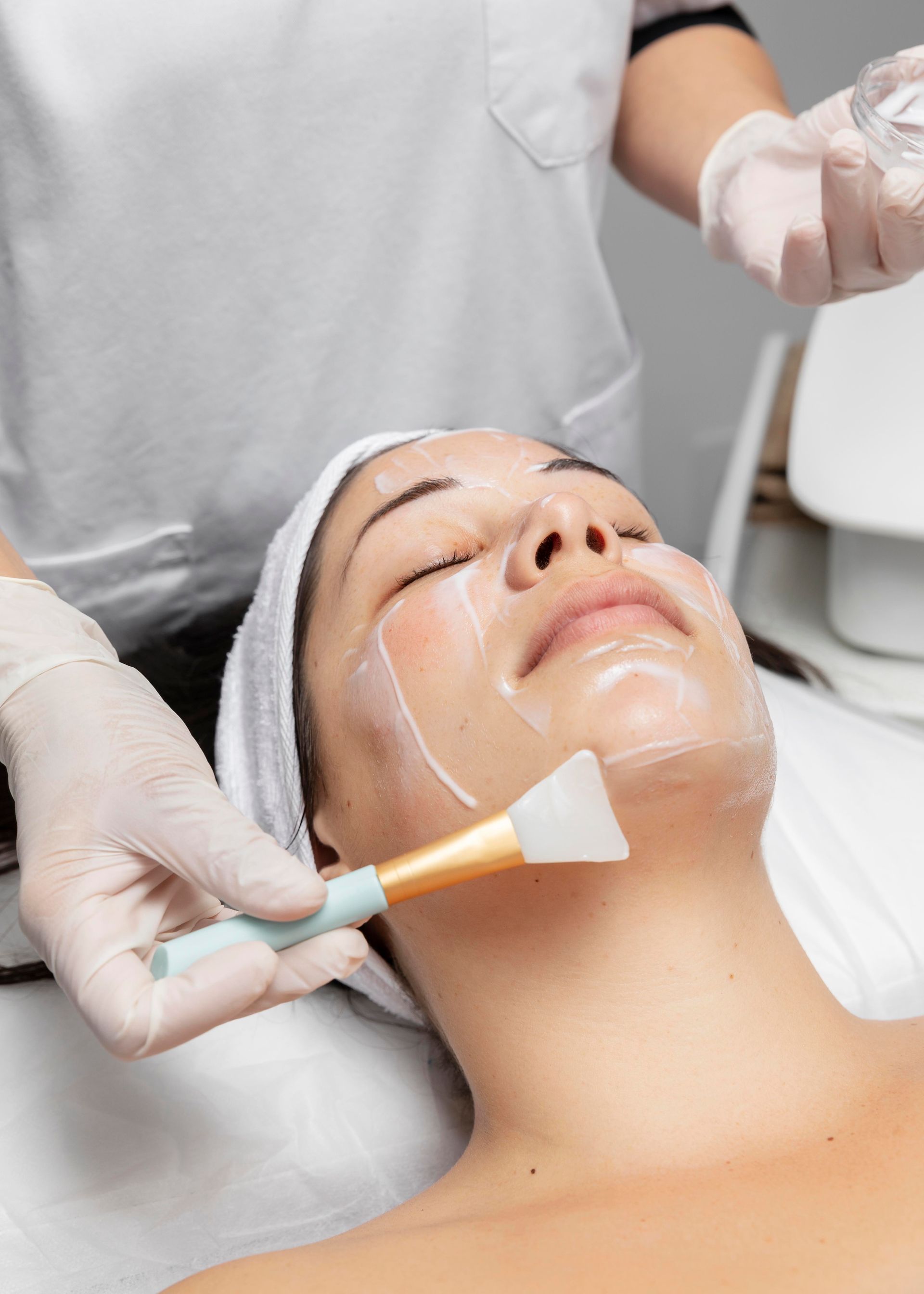 A spa professional wearing gloves applies a treatment mask to a client's face using a silicone brush.